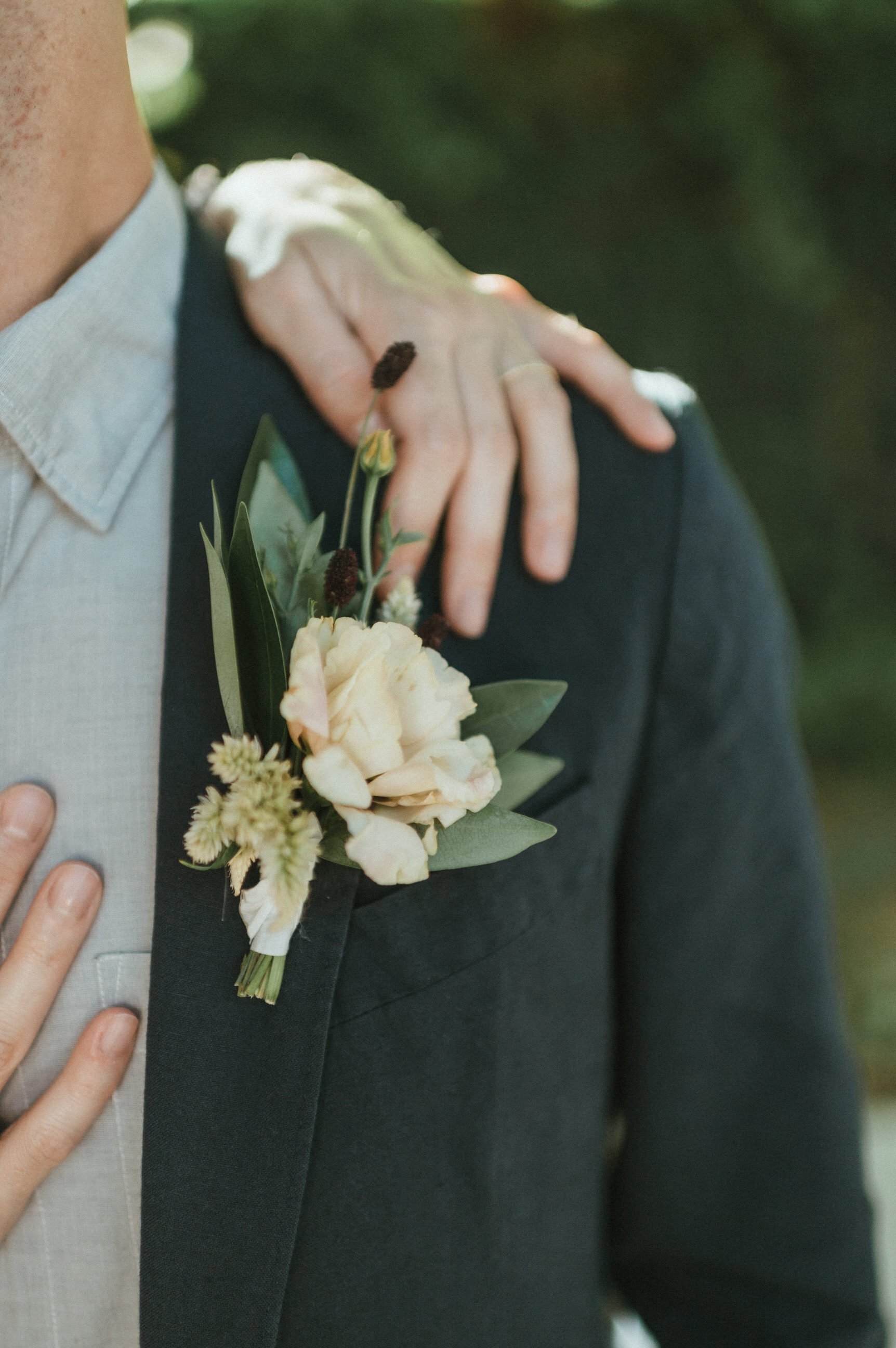 close up groom lapel with yellow flower and brides hand up over his shoulder near boutonniere