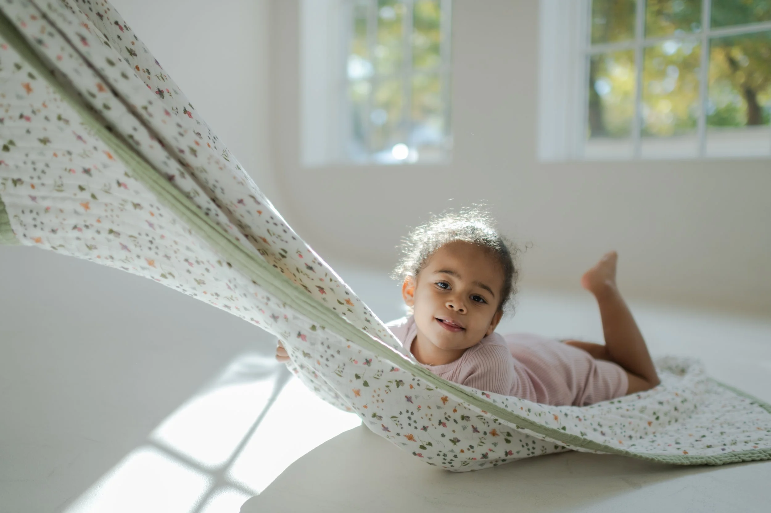 toddler with curly black hair riding on blanket while parent pulls it around in the kitchen