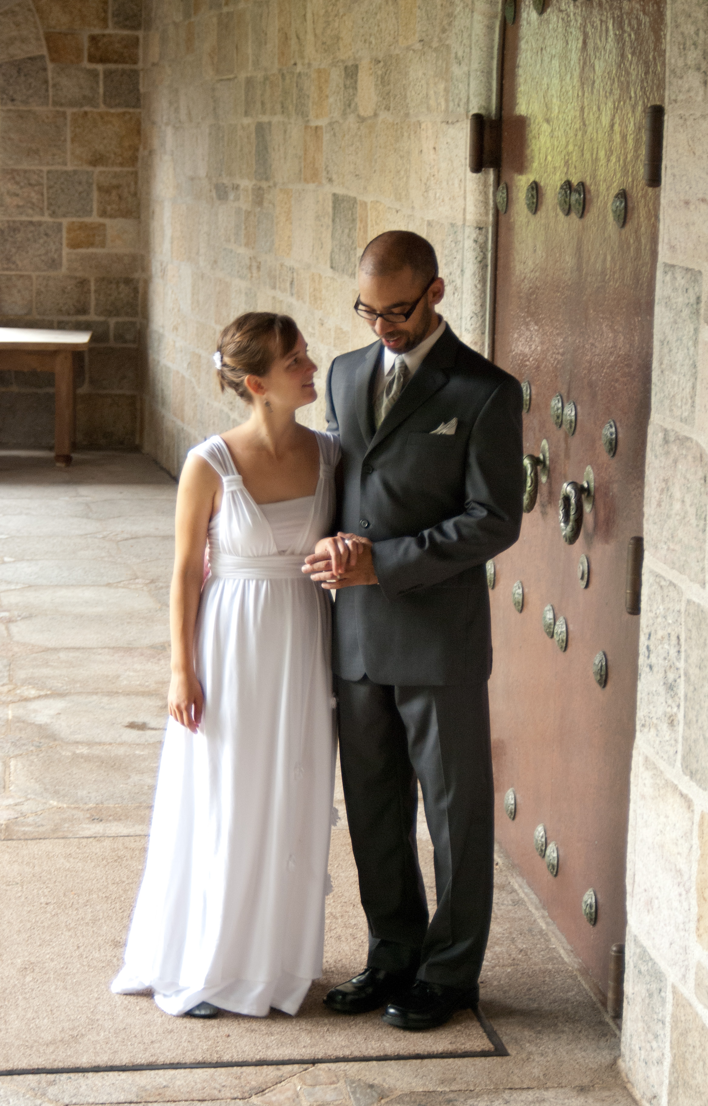The bride, in a white empire-waisted gown, holds hands with the groom, in a grey suit, between two columns in Glencairn Museum's cloister garden. 