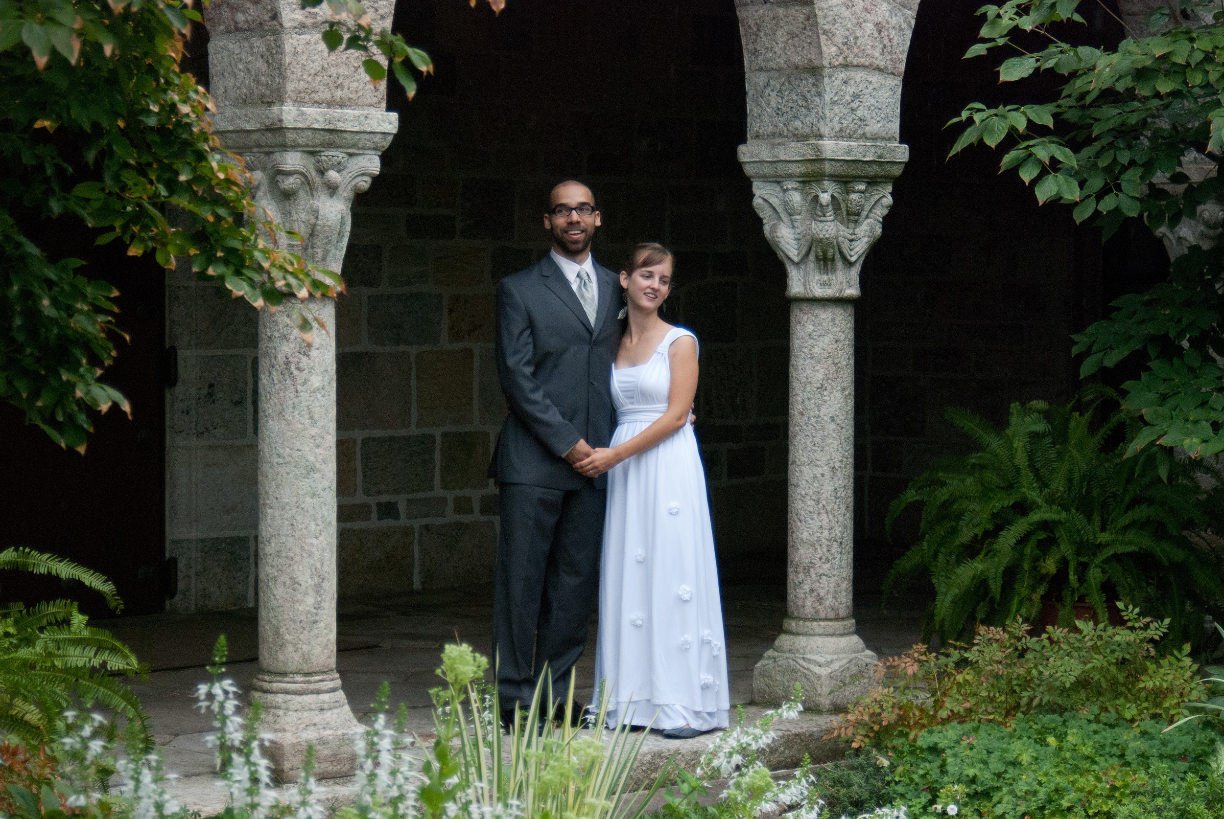 The bride, in a white empire-waisted gown, holds hands with the groom, in a grey suit, between two columns in Glencairn Museum's cloister garden. 