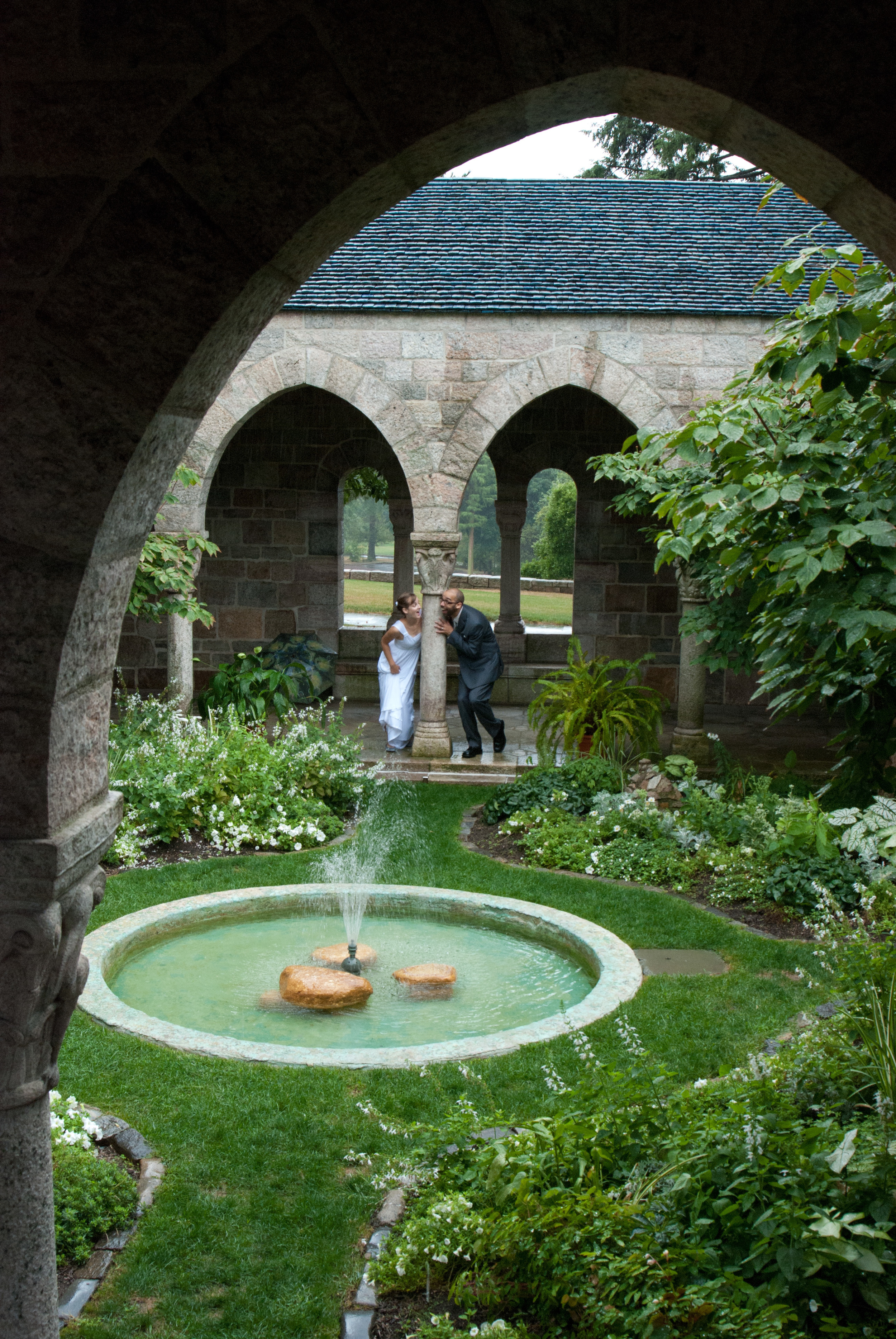 A bride and groom peek playfully at each other around a column in Glencairn Museum's cloister. The cloister fountain is in the foreground.