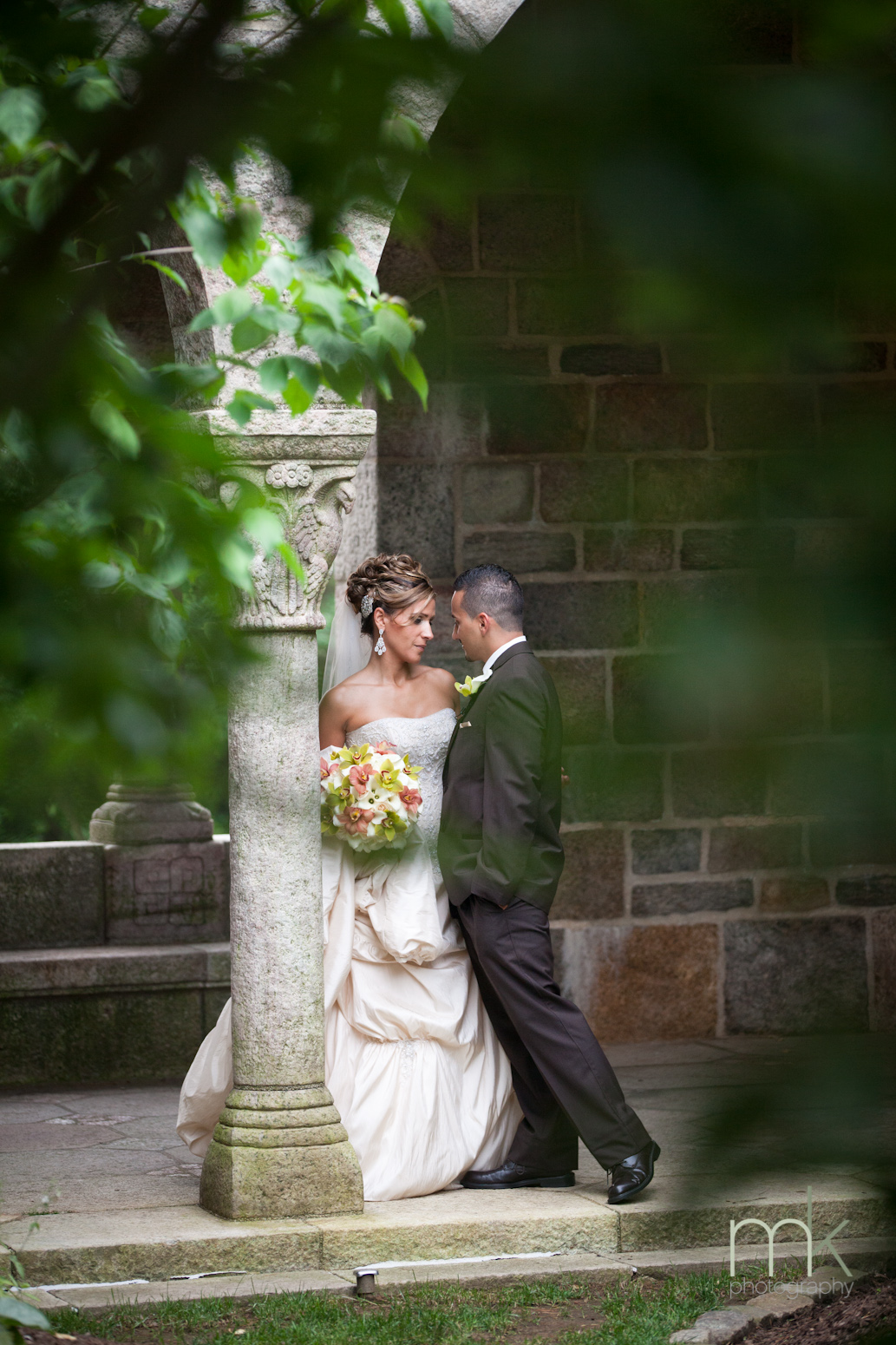 A bride in a white wedding dress and a groom in a black suit smile at each other while leaning against a column in Glencairn Museum's cloister.