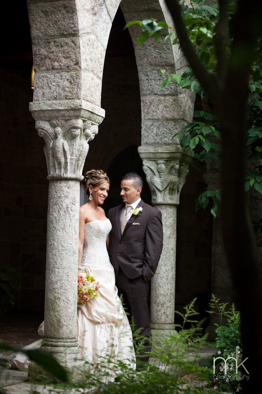 A bride in a white wedding gown leans against a column in Glencairn Museum's cloiser, while the groom in a dark suit stands next to her.