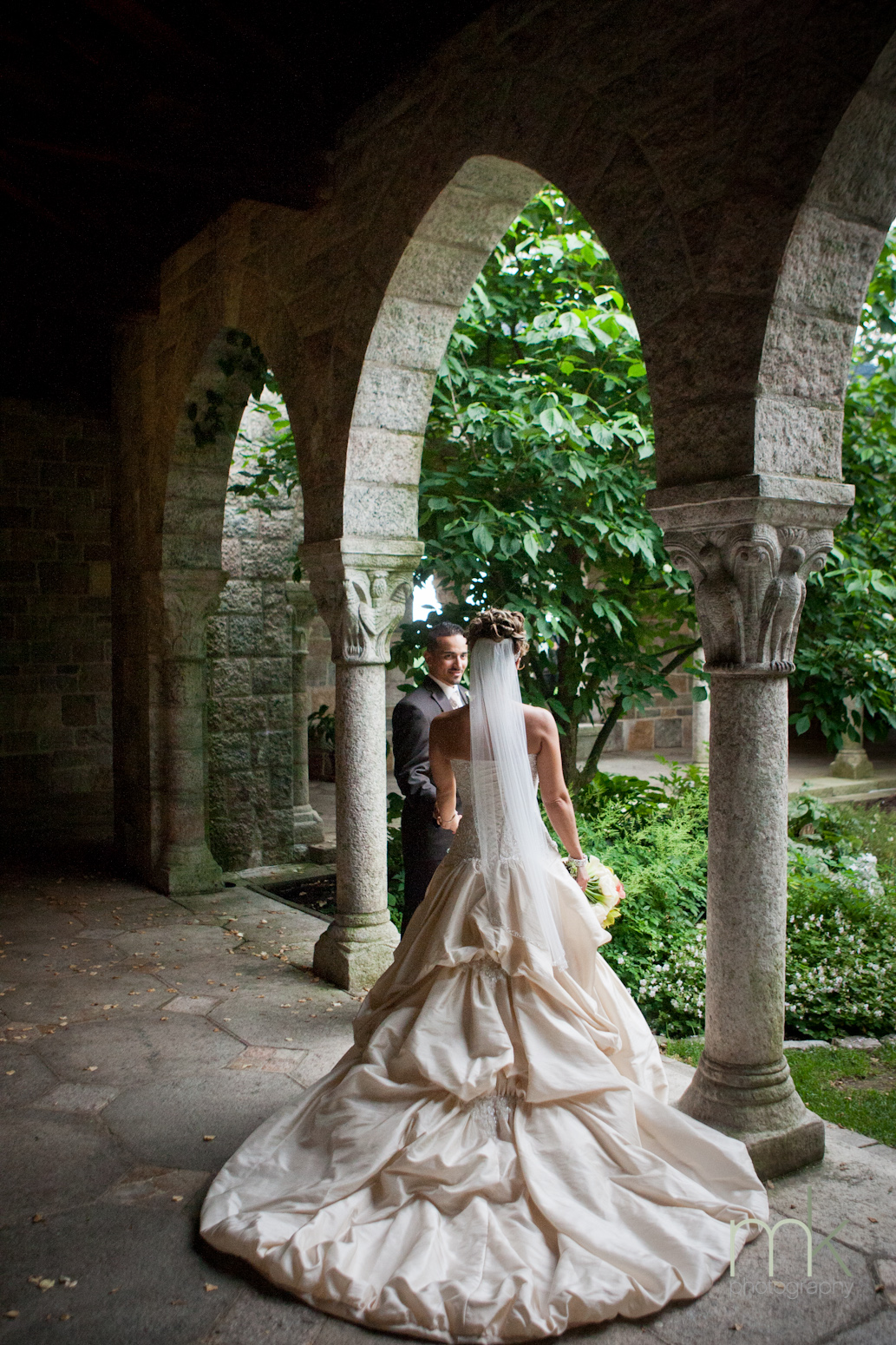 A groom stands facing the camera, and the bride stands with her back to the camera. Her ballgown style dress flows out behind her as they stand in Glencairn Museum's cloister.