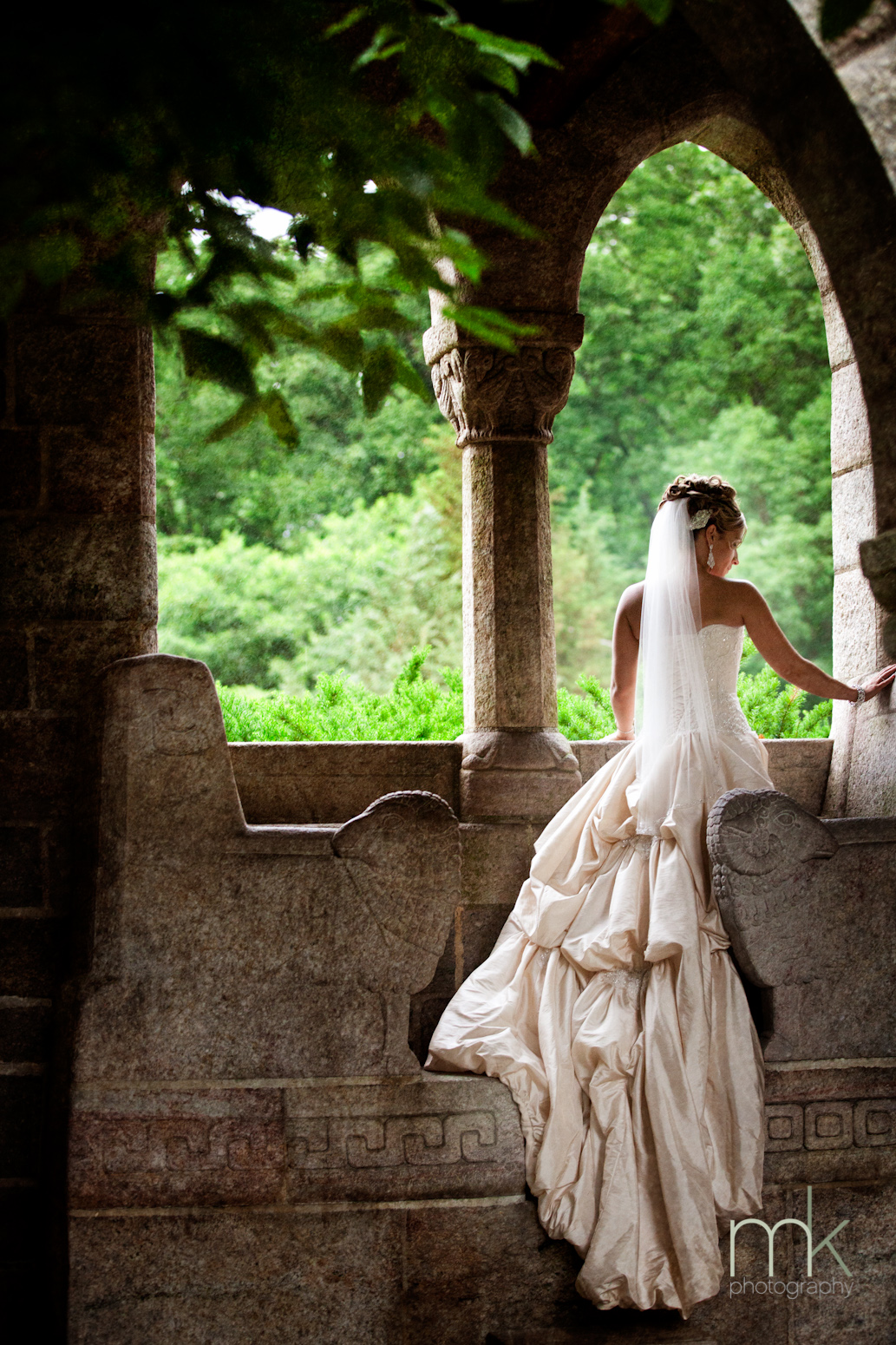 A bride in a ballgown dress stands on a stone bench in Glencairn Museum's cloister, with her back to the camera and her face in profile.