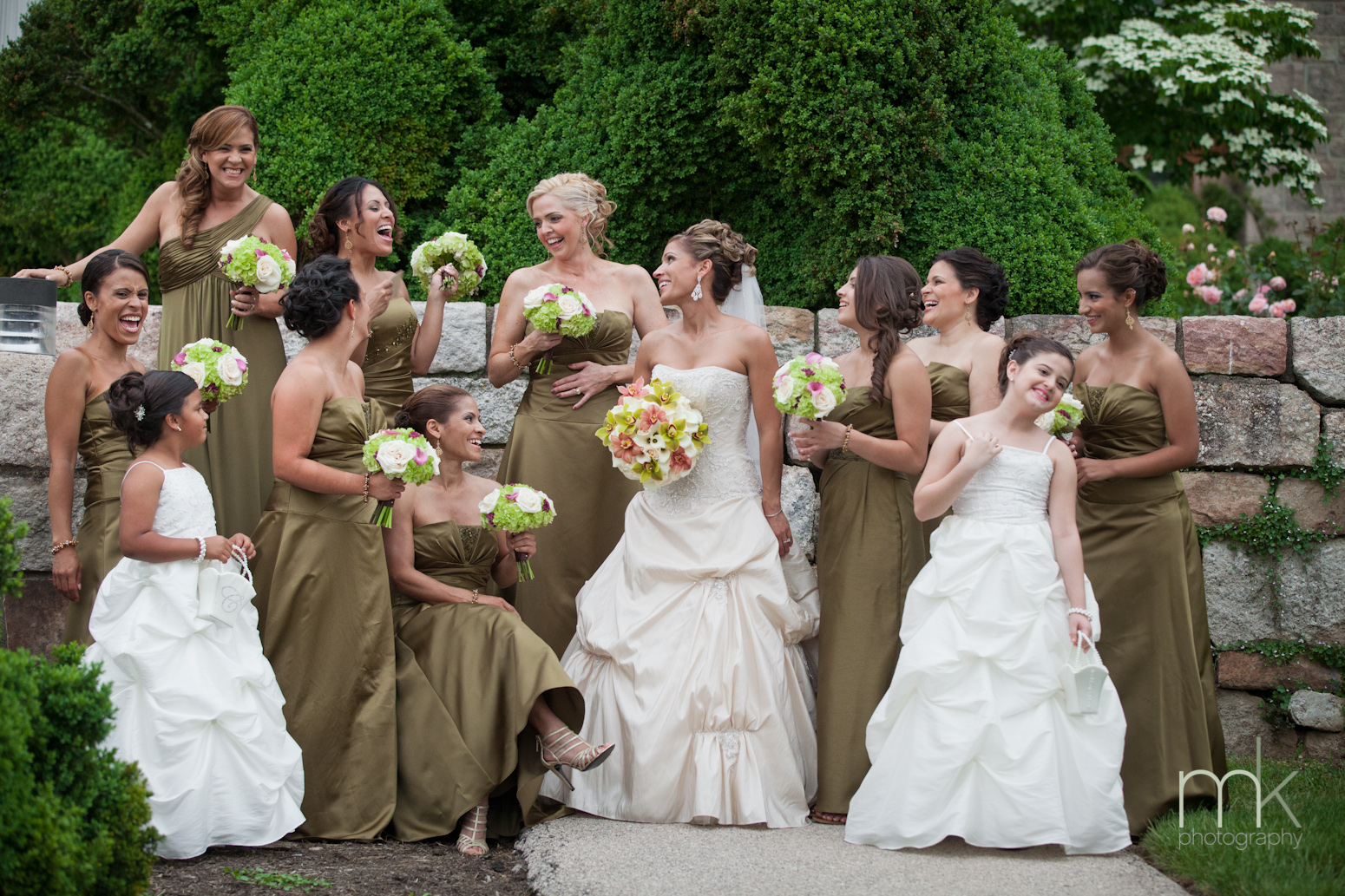 A bride in a ballgown with her bridesmaids in gold brown dresses and flower girls in white dresses smile and laugh by a stone wall outside Glencairn Museum.