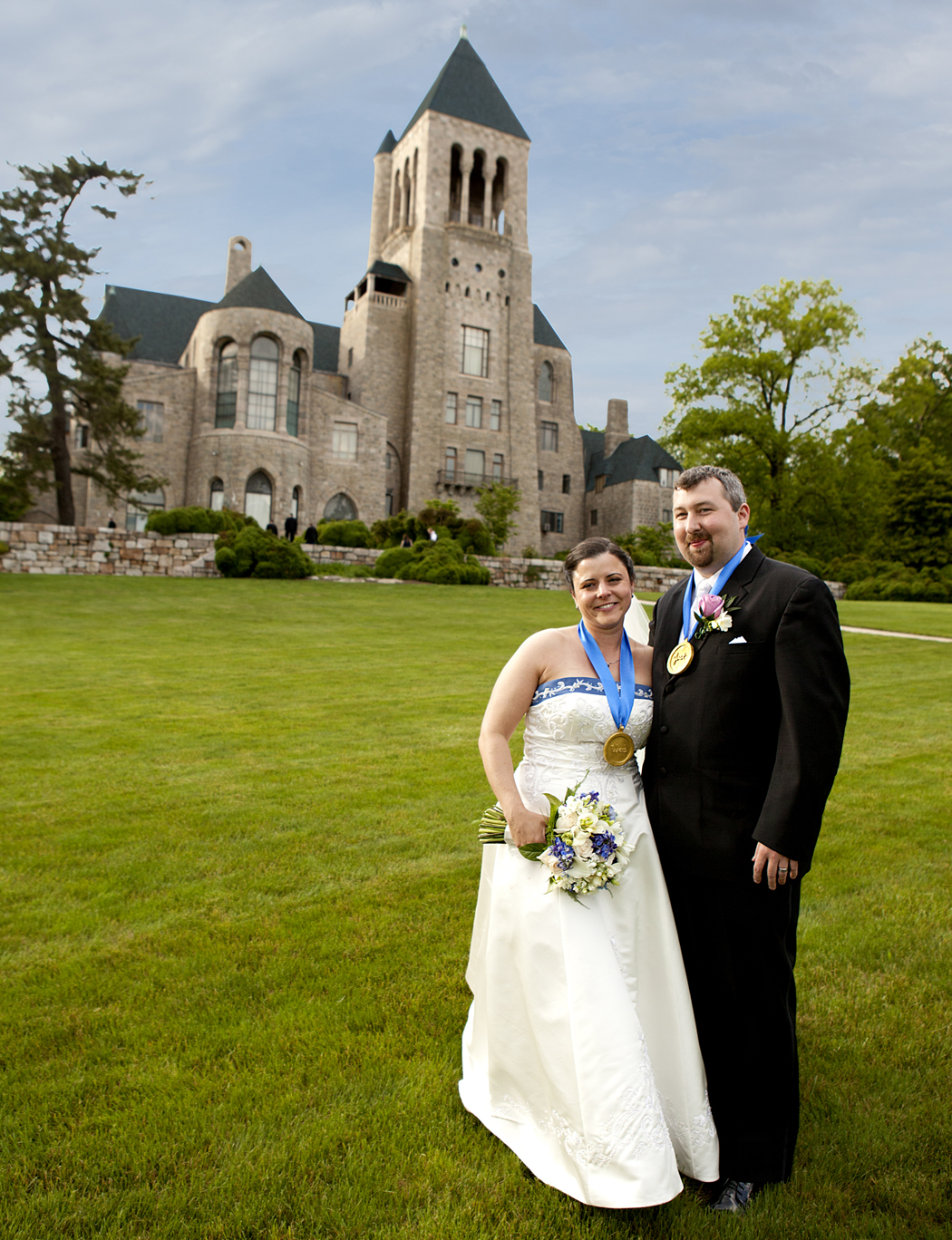 A bride and groom smile and stand hand-in-hand in a lawn, with Glencairn Museum in the background.