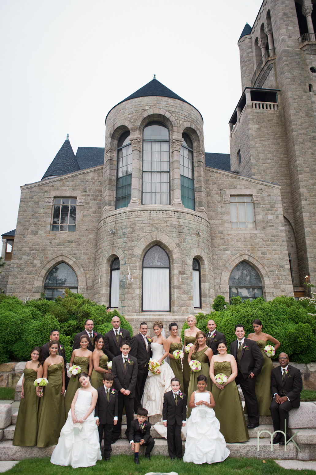 A large wedding party, the women in gold brown dresses, the flower girls in white dresses, and the men in dark suits, pose with the bride and groom in Glencairn Museum's sundial garden.