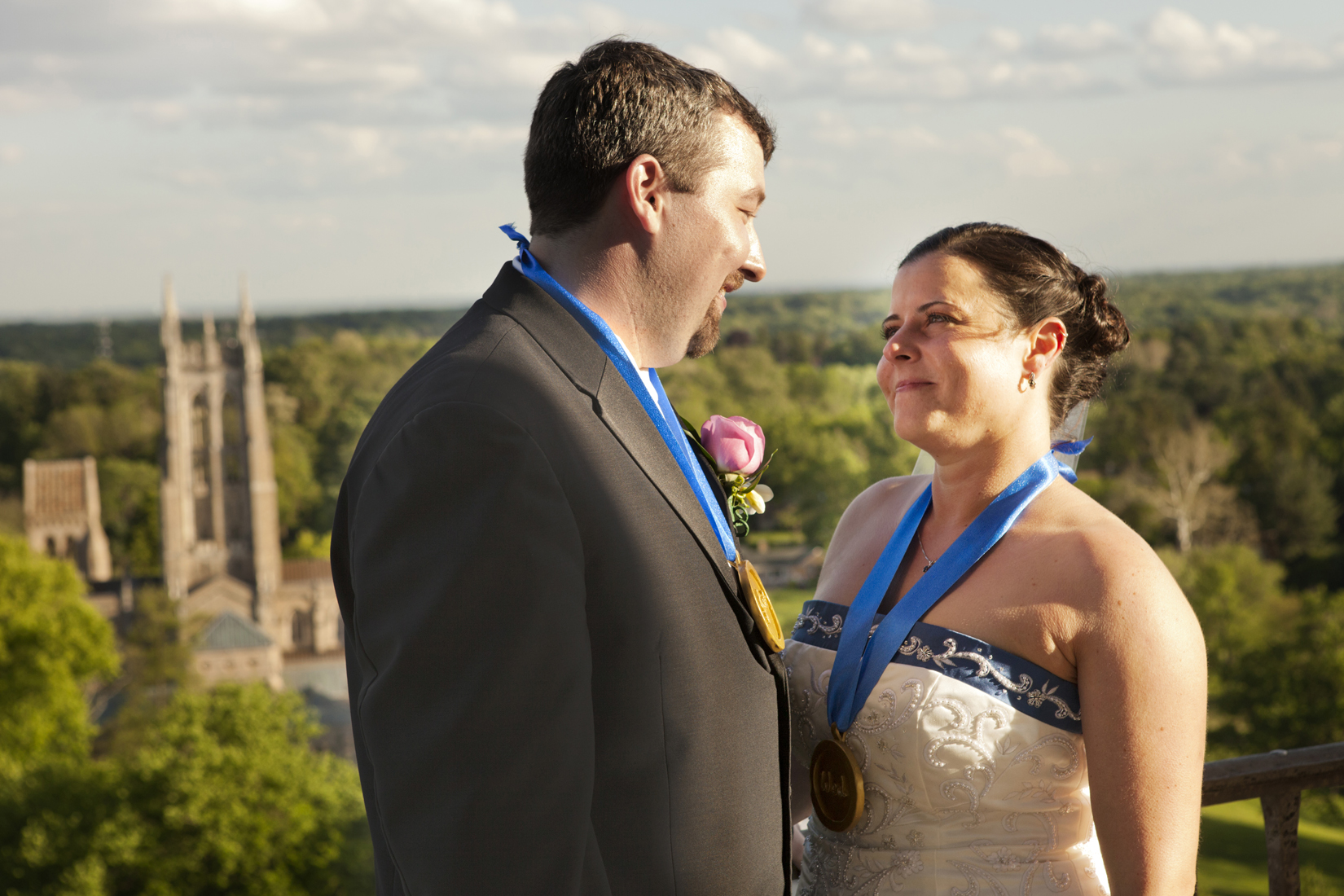 A bride and groom smile at each other while standing in Glencairn Museum's tower. The sunlit Bryn Athyn Cathedral is in the background.