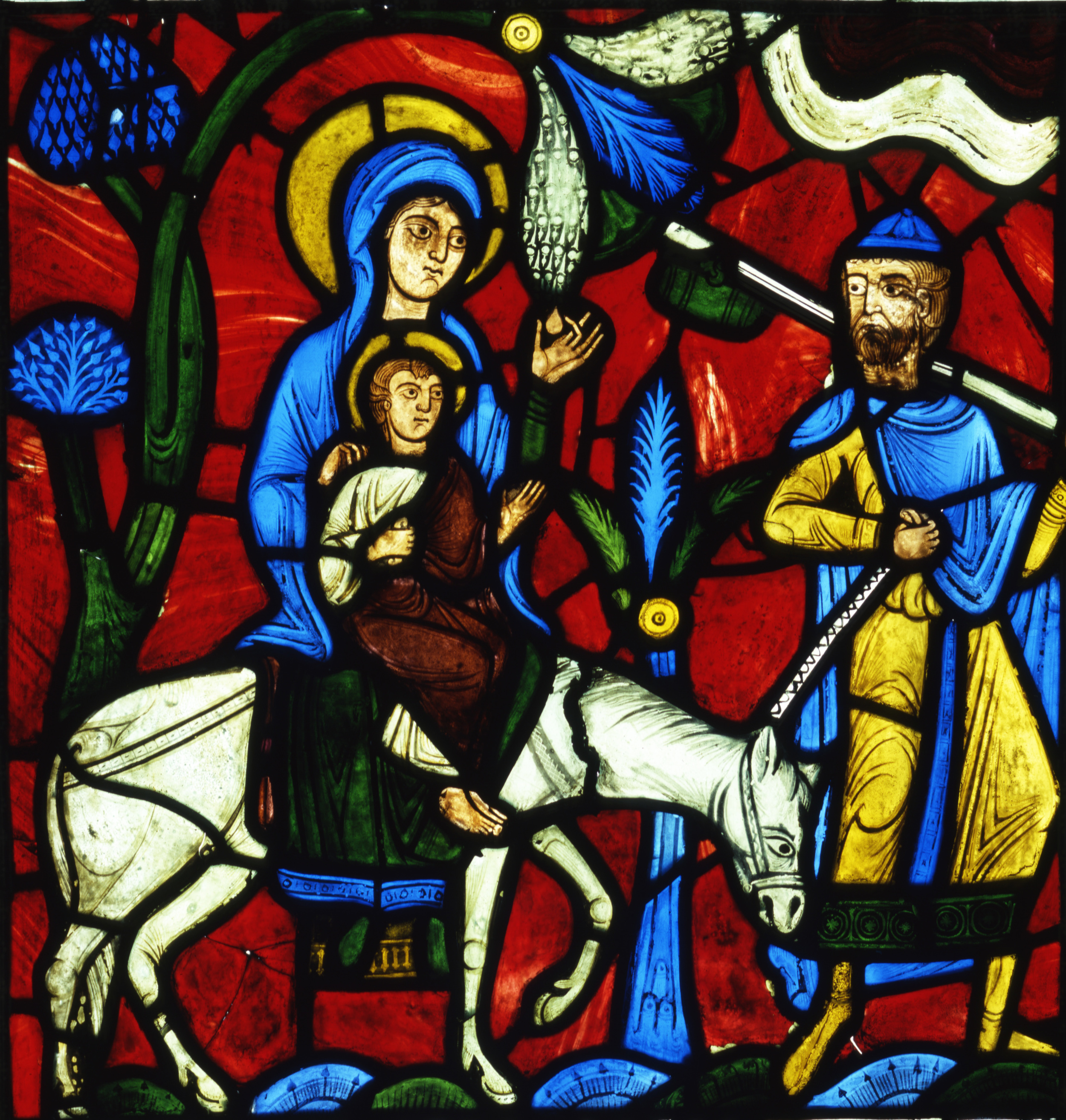 The Flight into Egypt, from the Infancy of Christ Window. Pot-metal glass. France, Abbey of Saint-Denis (near Paris), circa 1145. This stained glass panel depicts a legend surrounding the story of the Flight into Egypt. The biblical story from the Gospel of Matthew describes how Joseph was commanded in a dream to escape from Herod with Mary and the baby Jesus to Egypt. The legend from Pseudo-Matthew shown in this panel, not told in the Gospels, tells of a moment on the journey when Mary is weary and in need of food. Jesus commands the palm tree to bend down so his mother can pluck the fruit. Collection of Glencairn Museum (03.SG.114).