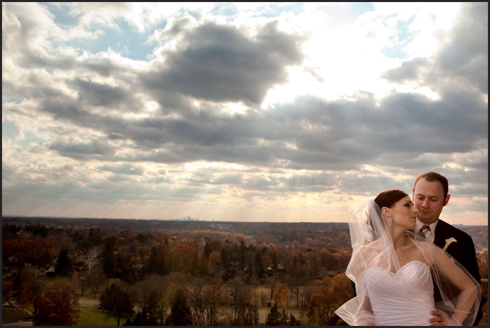 A bride and groom stand back to front, looking at each other, in Glencairn's tower. Behind them stretches a sunbeam-filled view from Glencairn Museum's tower, including the Philadelphia skyline.