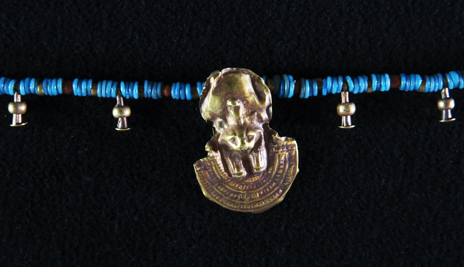 Figure 16: Aegis with Lioness . The larger object in this photo is an aegis , or shield, with a depiction of the head of a leonine goddess, probably Bastet or Tefnut, wearing a sundisk crown with a rearing uraeus. The bottom part of the aegis is a stylized form of a beaded wesekh (or “broad”) collar (see Figure 17). The aegis is a protective symbol, and this example was made by beating a thin sheet of metal (probably gold) into a mold. The detail on this amulet is exquisite. Glencairn Museum 15.JW.179.