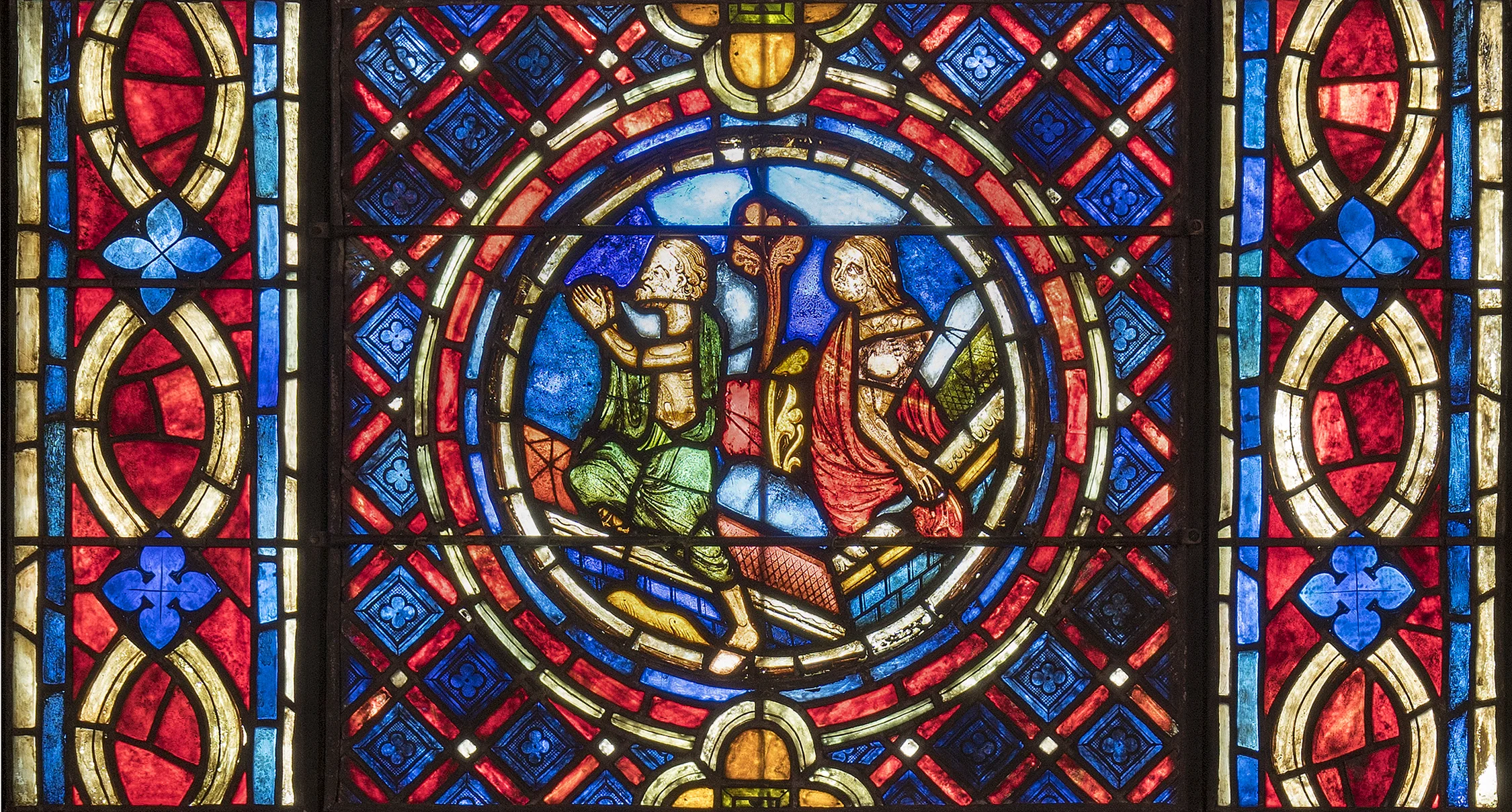 The Resurrected Rising: A man wearing a green burial shroud and a woman wearing a red shroud climb out of their tombs at the Last Judgment. The man is holding his hands in prayer. This thirteenth-century French stained glass roundel, in the collection of Glencairn Museum (ca. 1225, 03.SG.211), originally came from the east rose window of the parish church in Donne-marie-en-Montois (southwest of Provins). The roundel is now installed in one of the lancet windows on the east wall of Glencairn’s Great Hall.