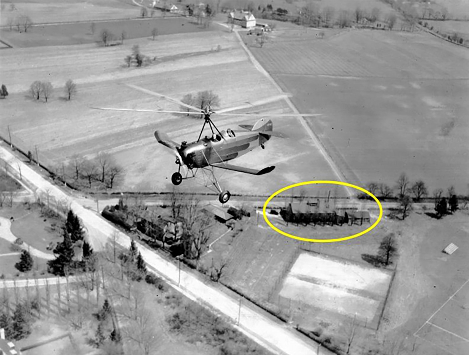 Figure 7: A Pitcairn PAA-1 Sport autogiro with one passenger flies over the Bryn Athyn glass factory (circled) and the old Academy of the New Church tennis courts. The Huntingdon Turnpike, the main thoroughfare, is clearly visible, as is the hay barn (top) for Cairnwood Farms. The glassworks made its first batch of glass on July 5, 1922, and was in continuous operation until April, 1942.