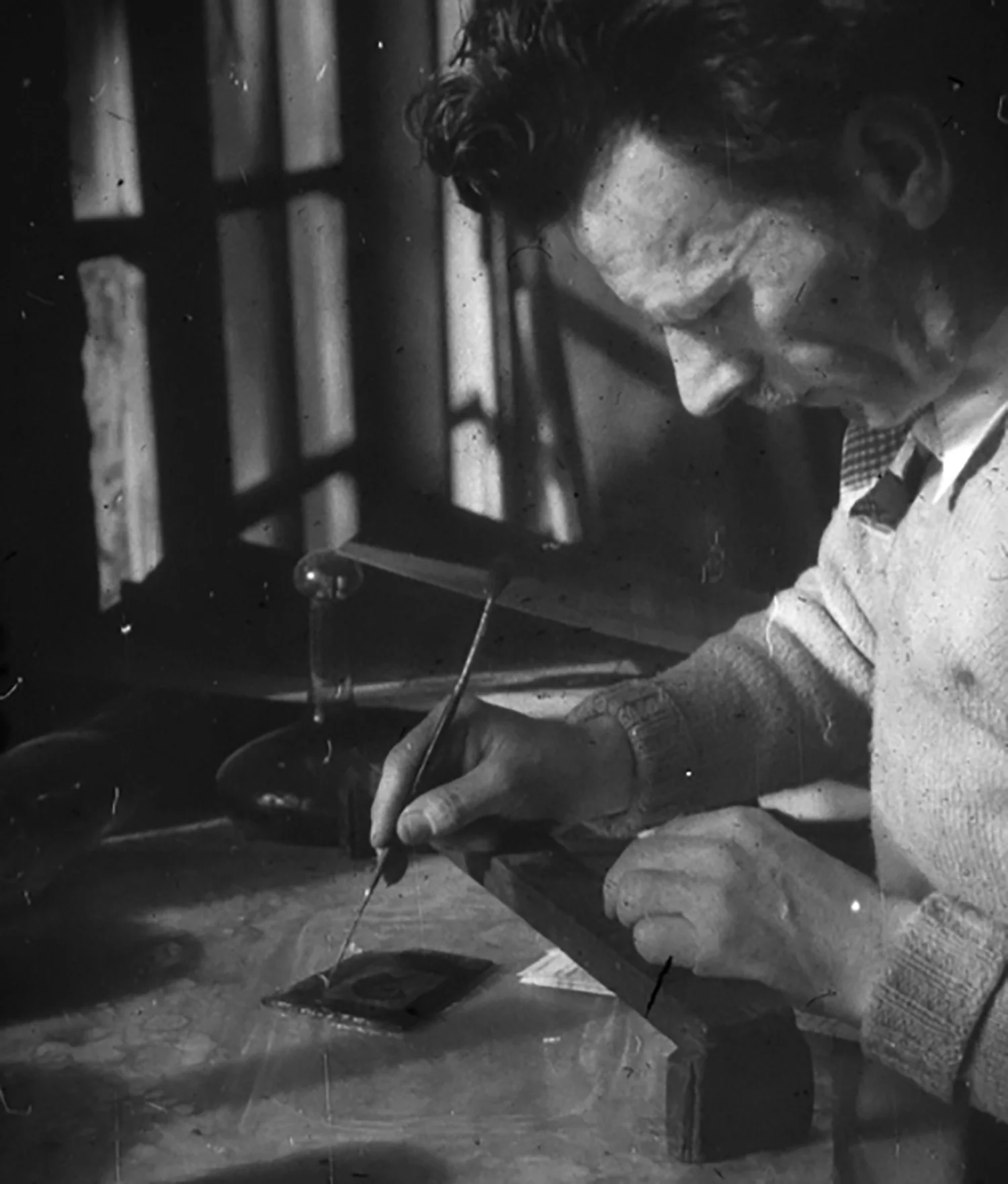 Figure 15: Once the pieces of glass were cut, the Bryn Athyn artists carefully painted on the faces, hands, feet, and other details. The pieces were then fired in a small electric kiln, bonding the paint to the glass. In this photograph, Albert E. Cullen paints a piece of glass using a “bridge” to steady his hand.