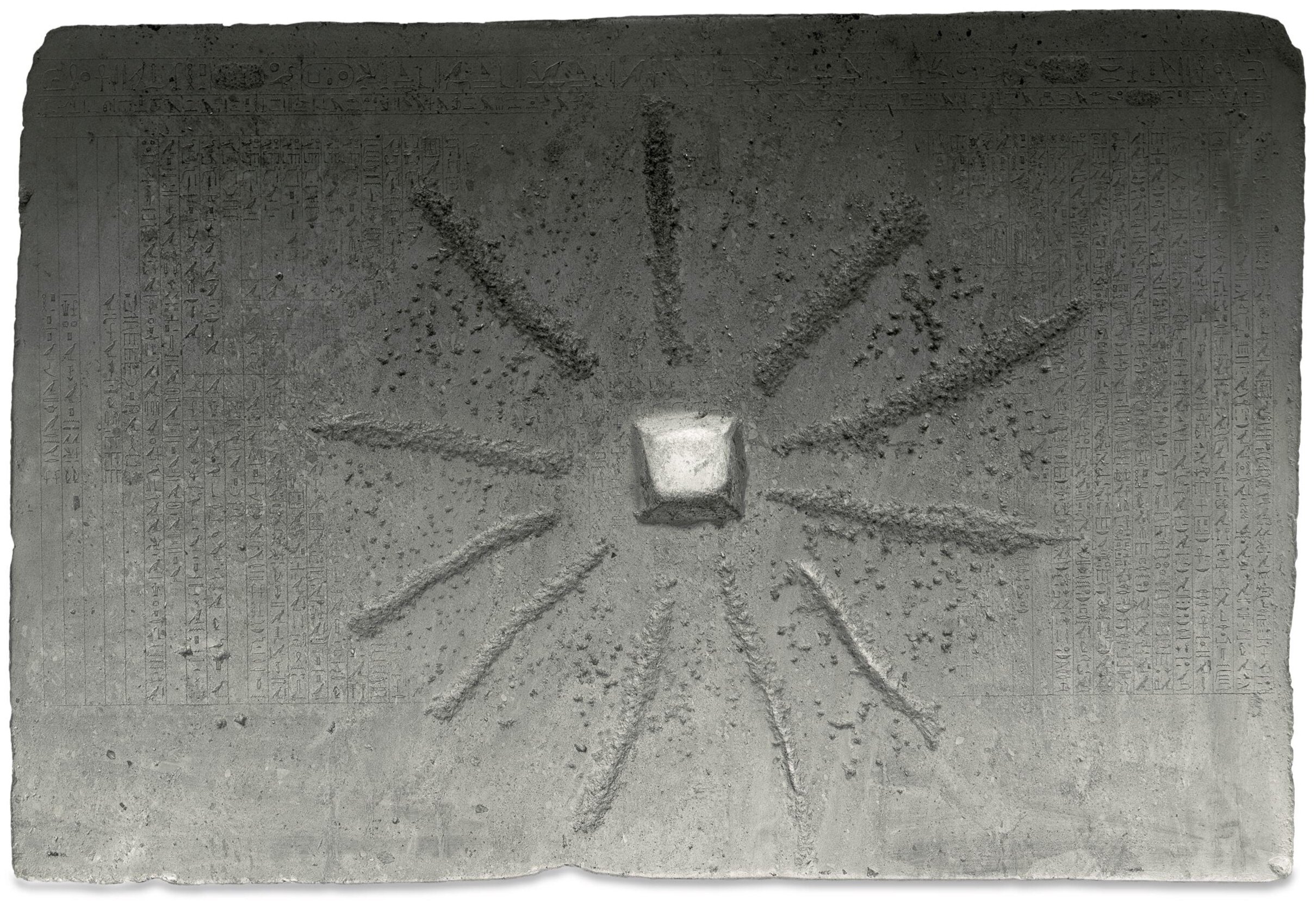 A grey rectangle stone, covered in hieroglyphic text. Eleven mostly straight carved lines radiate out from a square divot in the center, obscuring some of the text.