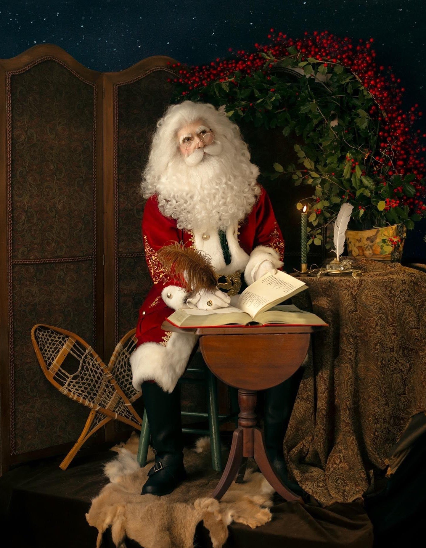  Santa poses with his book of names in a cozy space with a candle, fur skin rug, snow shoes, a vase of holly, and a starry sky above. 