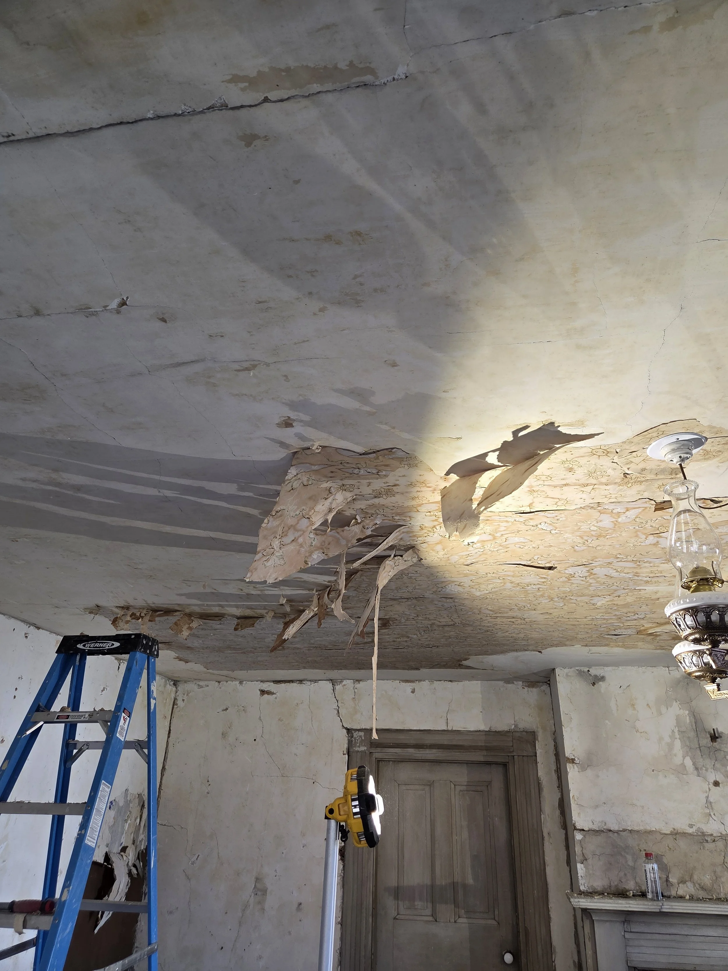 A ceiling with peeling wallpaper and exposed plaster, with a ladder and a work light set up underneath.