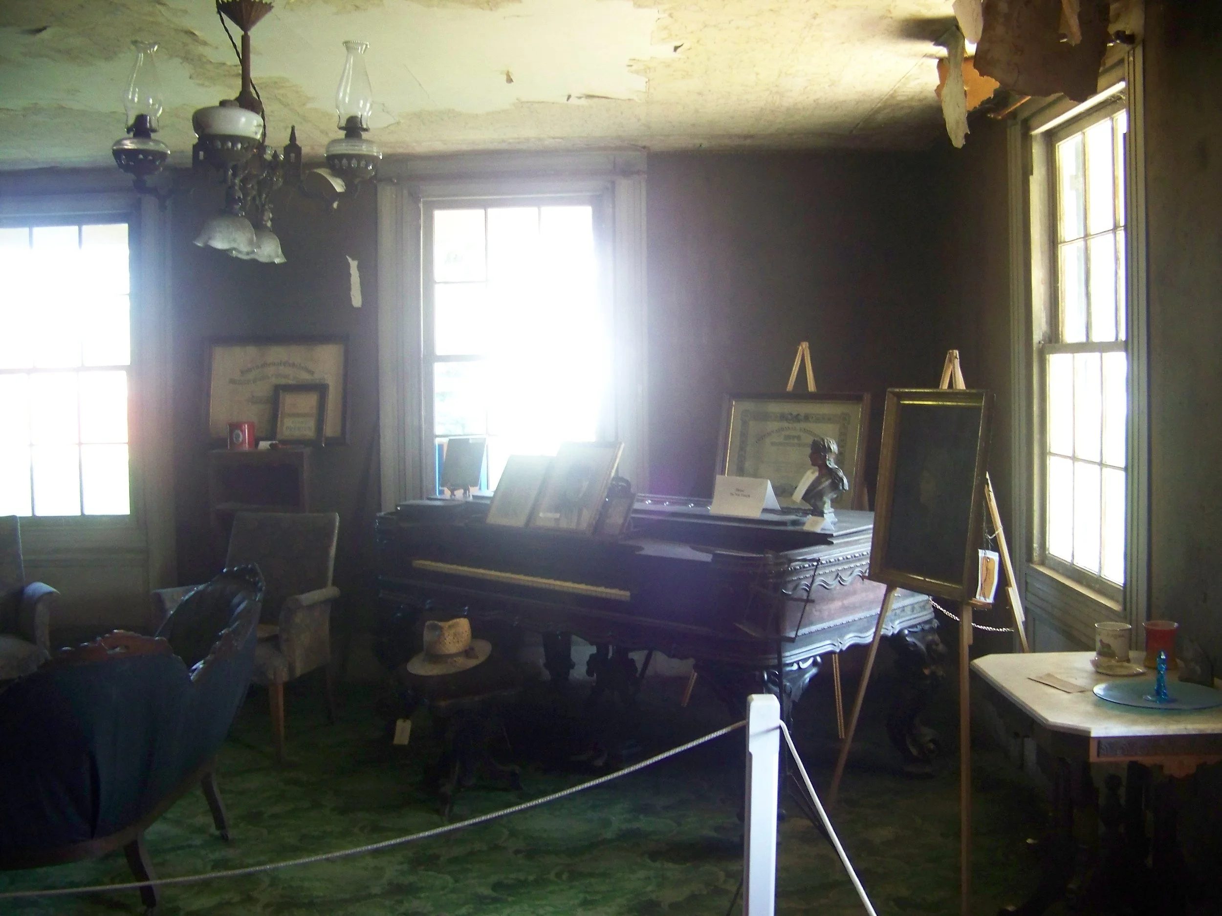 Old, dimly lit room with a grand piano, framed pictures and artwork, vintage furniture, a chandelier, and large windows letting in sunlight.
