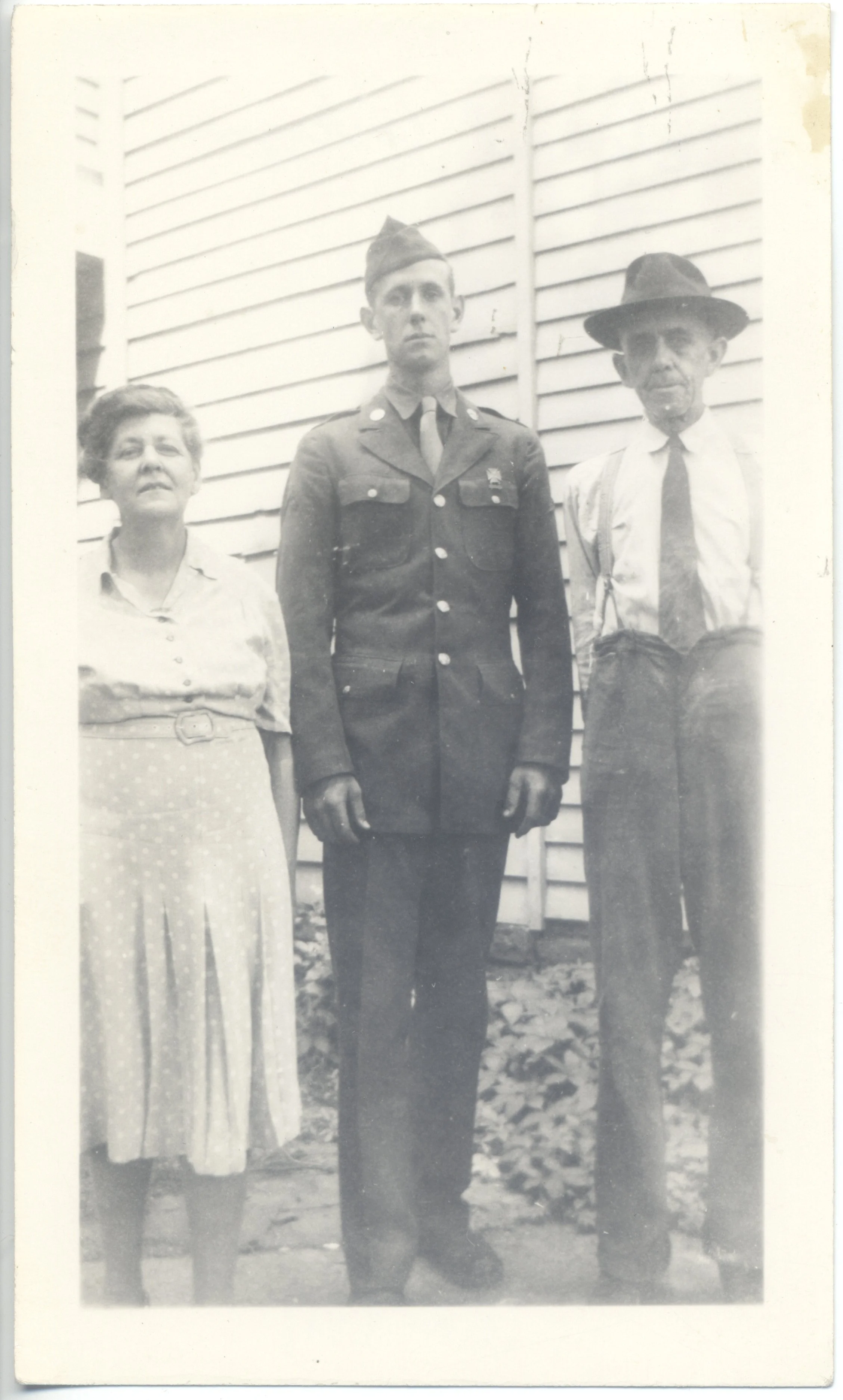 Black and white photograph of three people standing outdoors, with a woman on the left, a young man in a military uniform in the middle, and an older man on the right, against a building with horizontal siding.