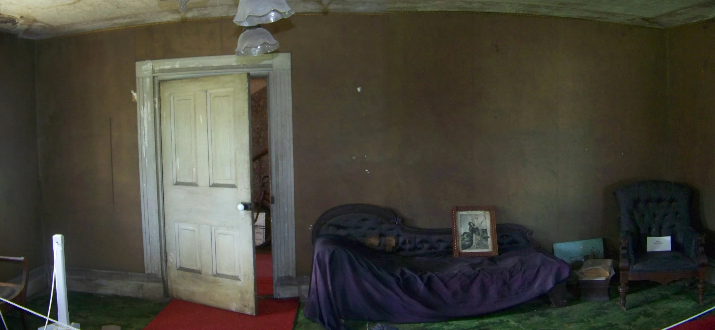 An old room with brown walls, a white door slightly open revealing a staircase, and vintage furniture including a sofa covered with a purple cloth, a framed picture resting on the sofa, and a dark armchair. A red carpet is on a green floor.