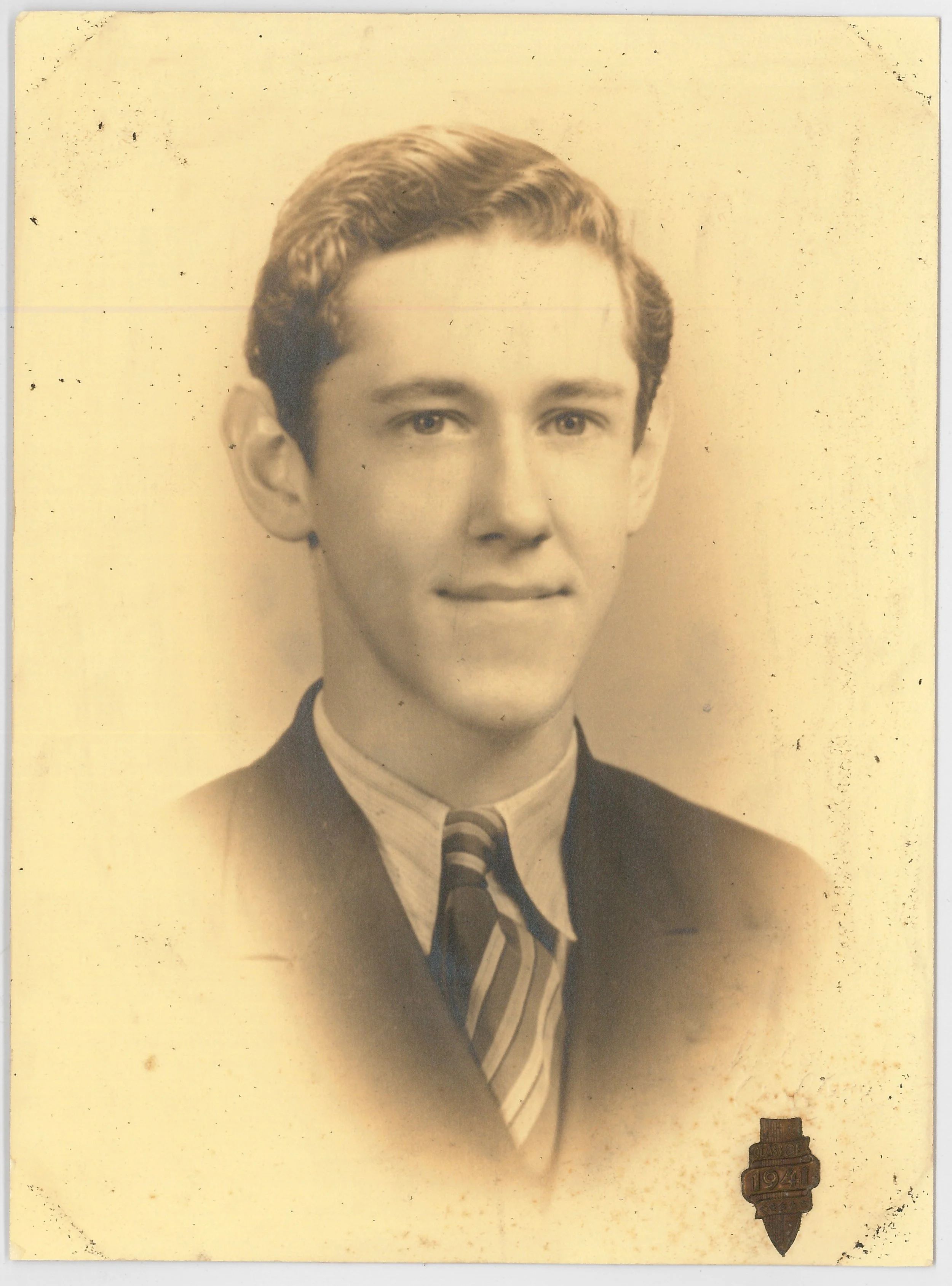 A vintage black and white portrait of a young man in a suit, tie, and collared shirt, with a badge or pin on his lapel, likely from 1941.