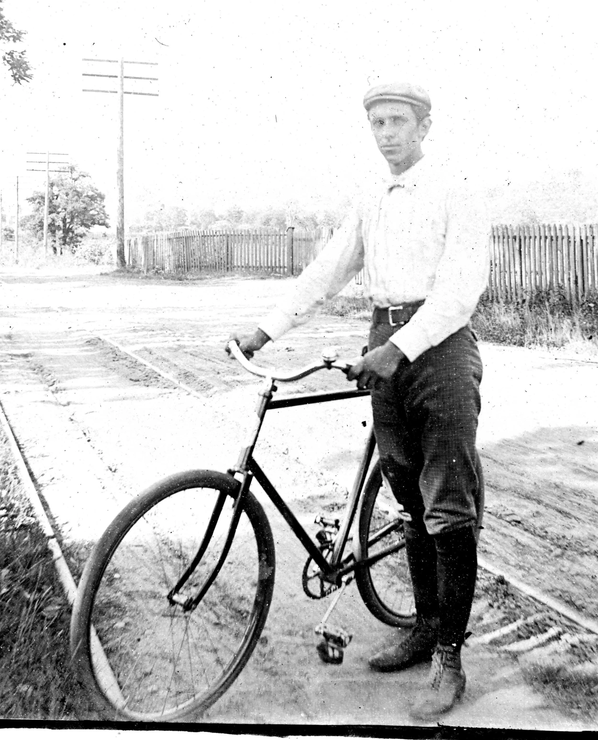 A young Zan in vintage clothing standing next to a bicycle on a dirt road, with wooden fences and utility poles in the background.