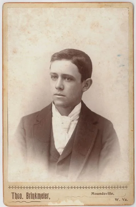 Black and white photograph of a young man in formal attire, including a dark jacket, white shirt, and vest, with short dark hair, posing against a plain background. The photo has a decorative border with the name "Théo. Brinkmeier" and location "Moundsville, W. Va." at the bottom.