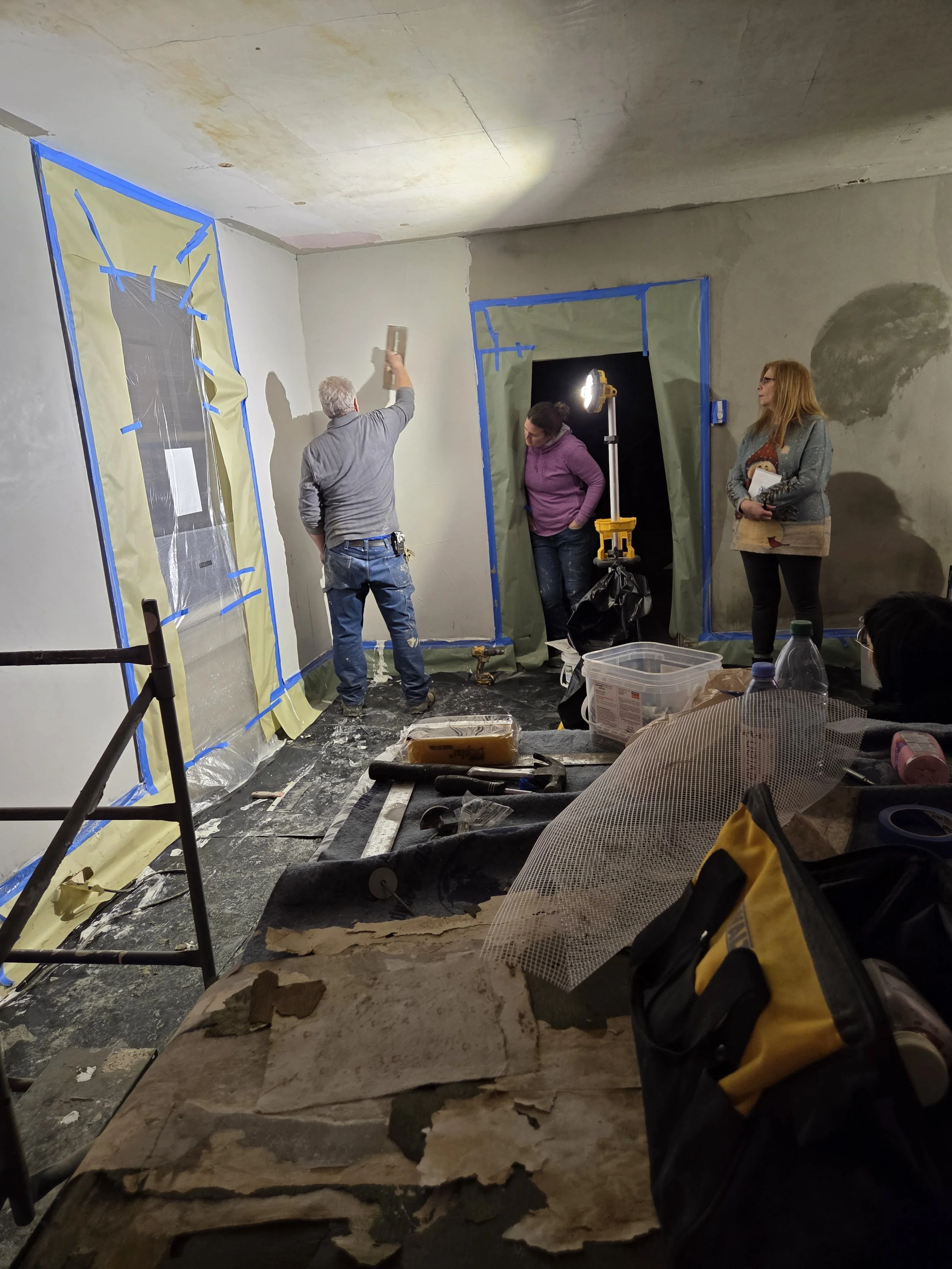 People working on renovating interior walls in a room, with blue painter's tape and protective coverings on the windows, construction tools, and materials scattered around.