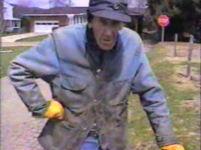 Sam Cockayne wearing a gray jacket and a cap, leaning forward outdoors on a sidewalk, with a residential street in the background.