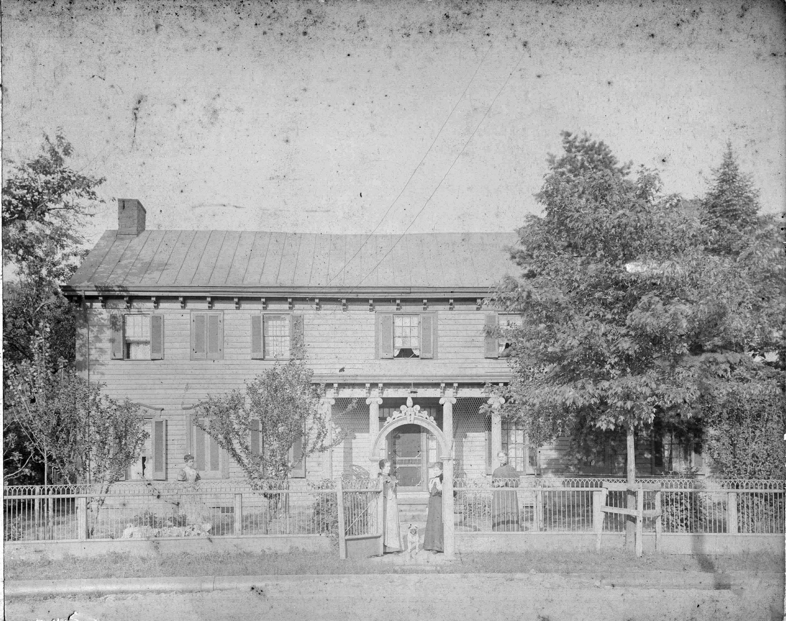 Old black-and-white photo of Valley Farmstead a large two-story house with eight shuttered windows, a front porch, and a metal roof. The house is surrounded by trees and a picket fence, with three women and two children standing in front of the gate.