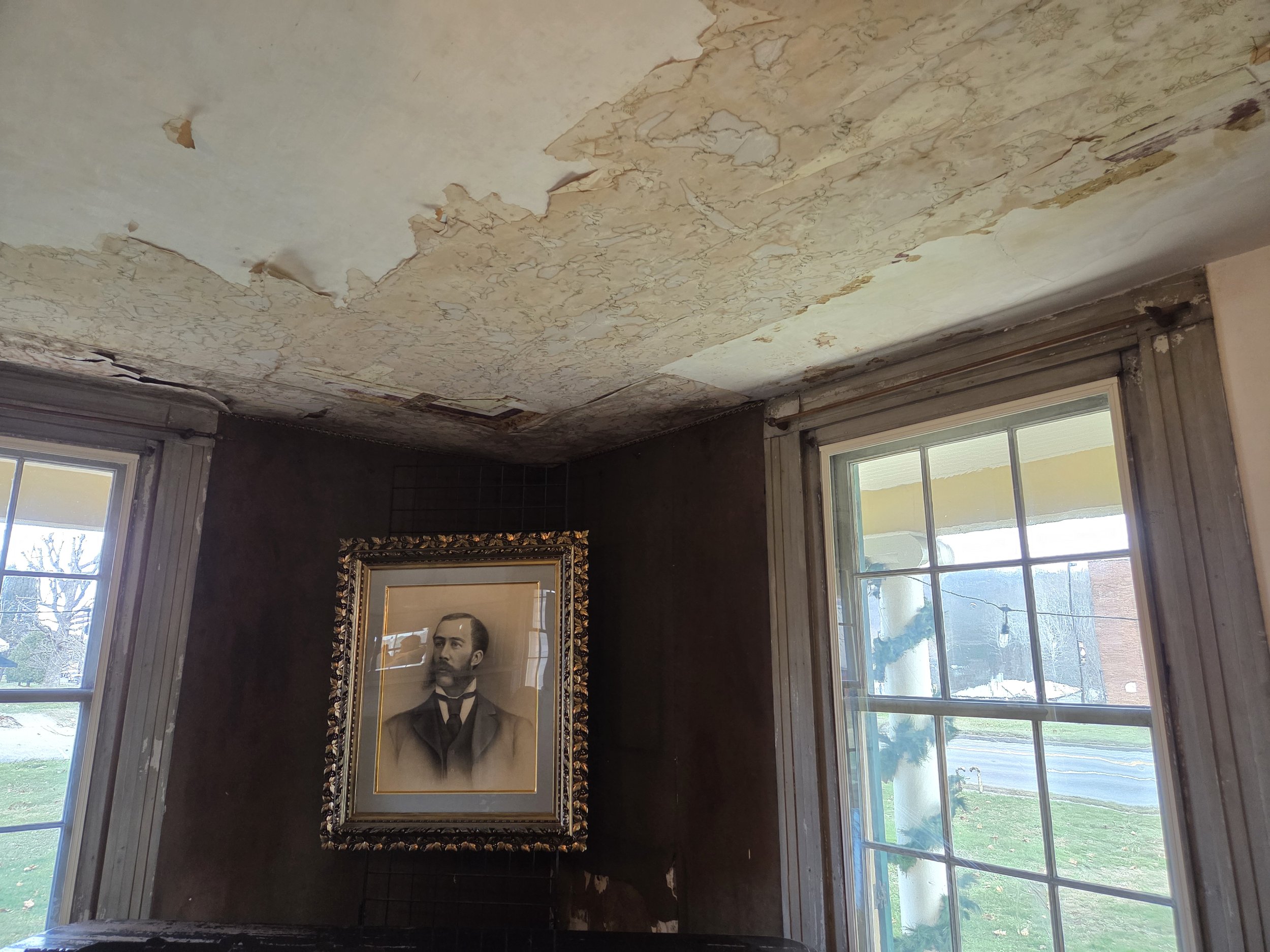 A room with peeling wallpaper on the ceiling, two large windows with a view of a street outside, and a framed portrait of a man with a beard and suit hanging on the wall.