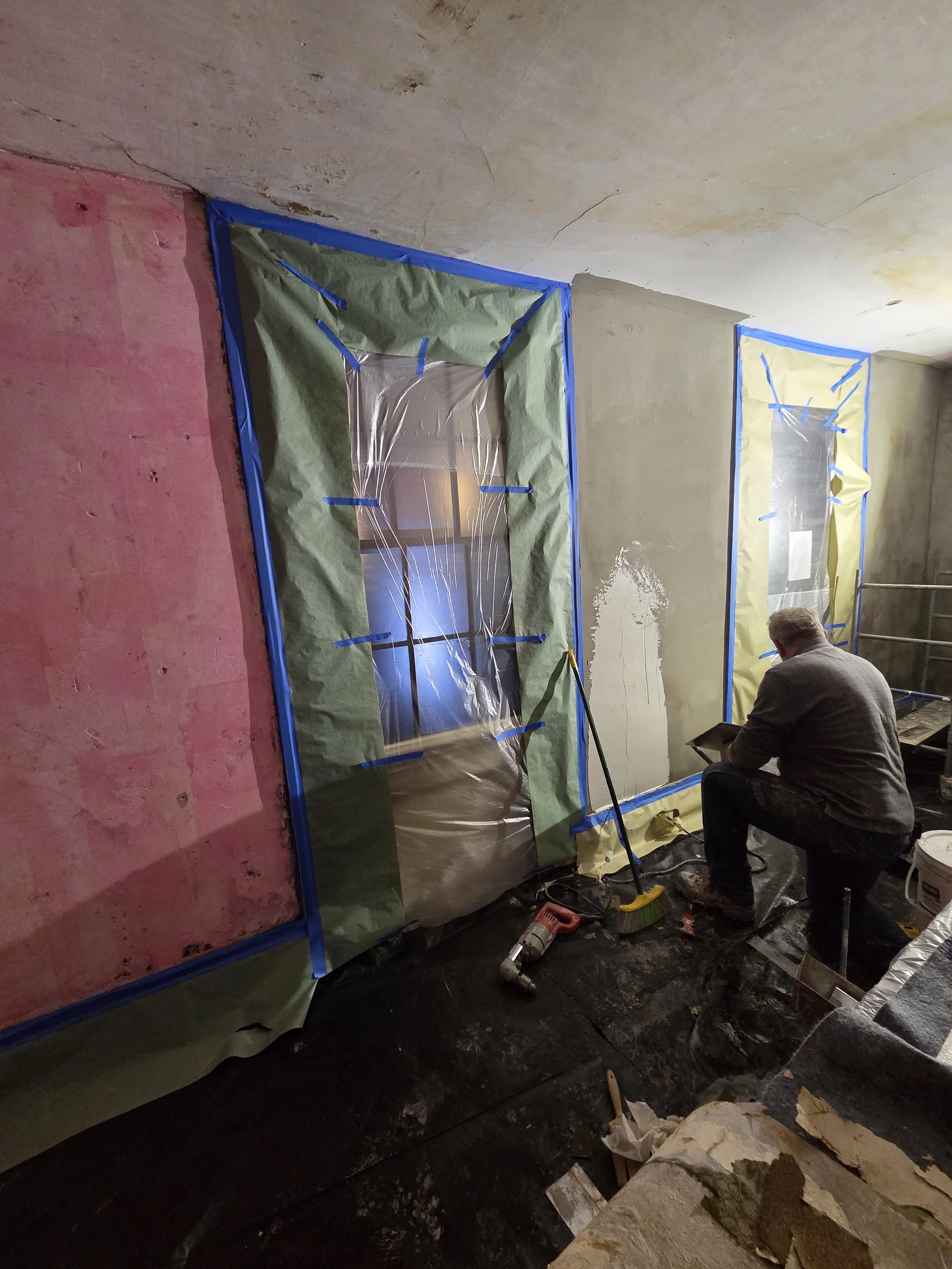 Person working on a construction or renovation project on interior wall, with windows covered in plastic and tape, surrounding a window.