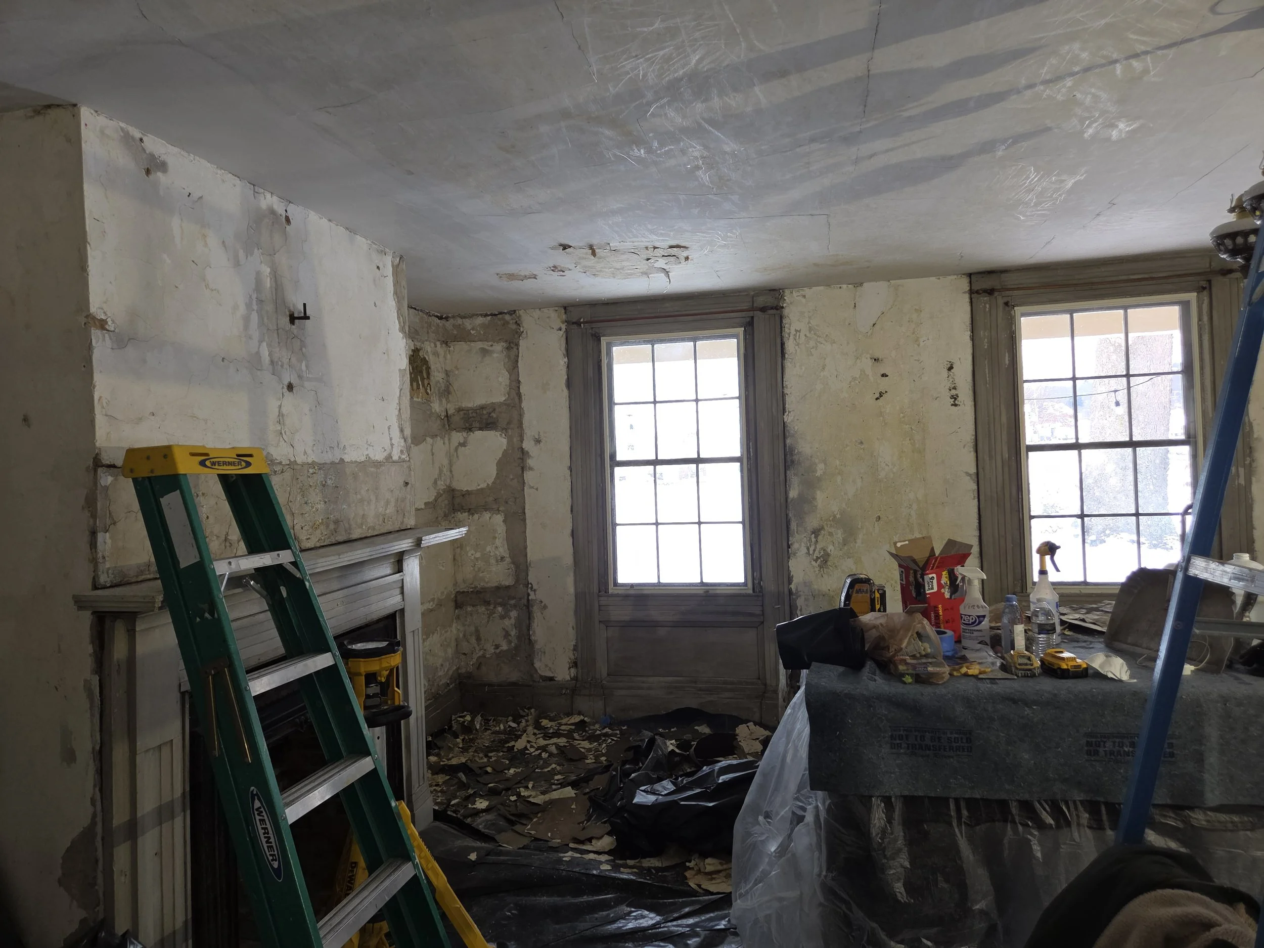 A room under renovation with peeling paint on the walls, construction tools and supplies on the table, a green ladder, and two windows letting in natural light.