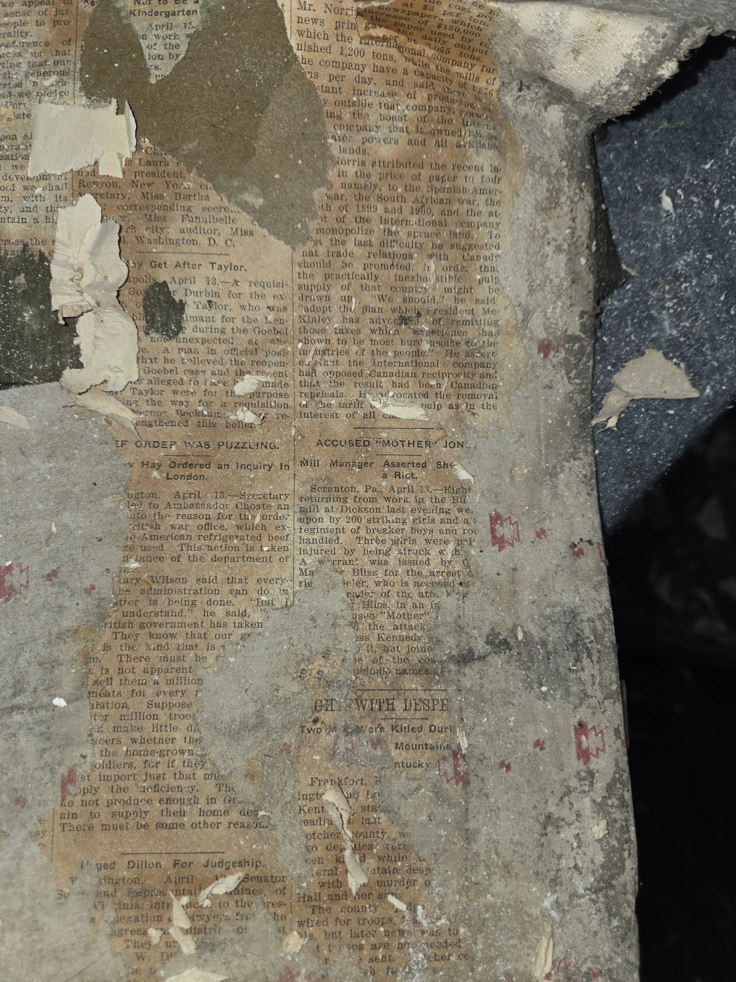 A torn piece of newspaper with peeling edges, embedded in a rough, cracked surface with bits of dried paint and ink stains.