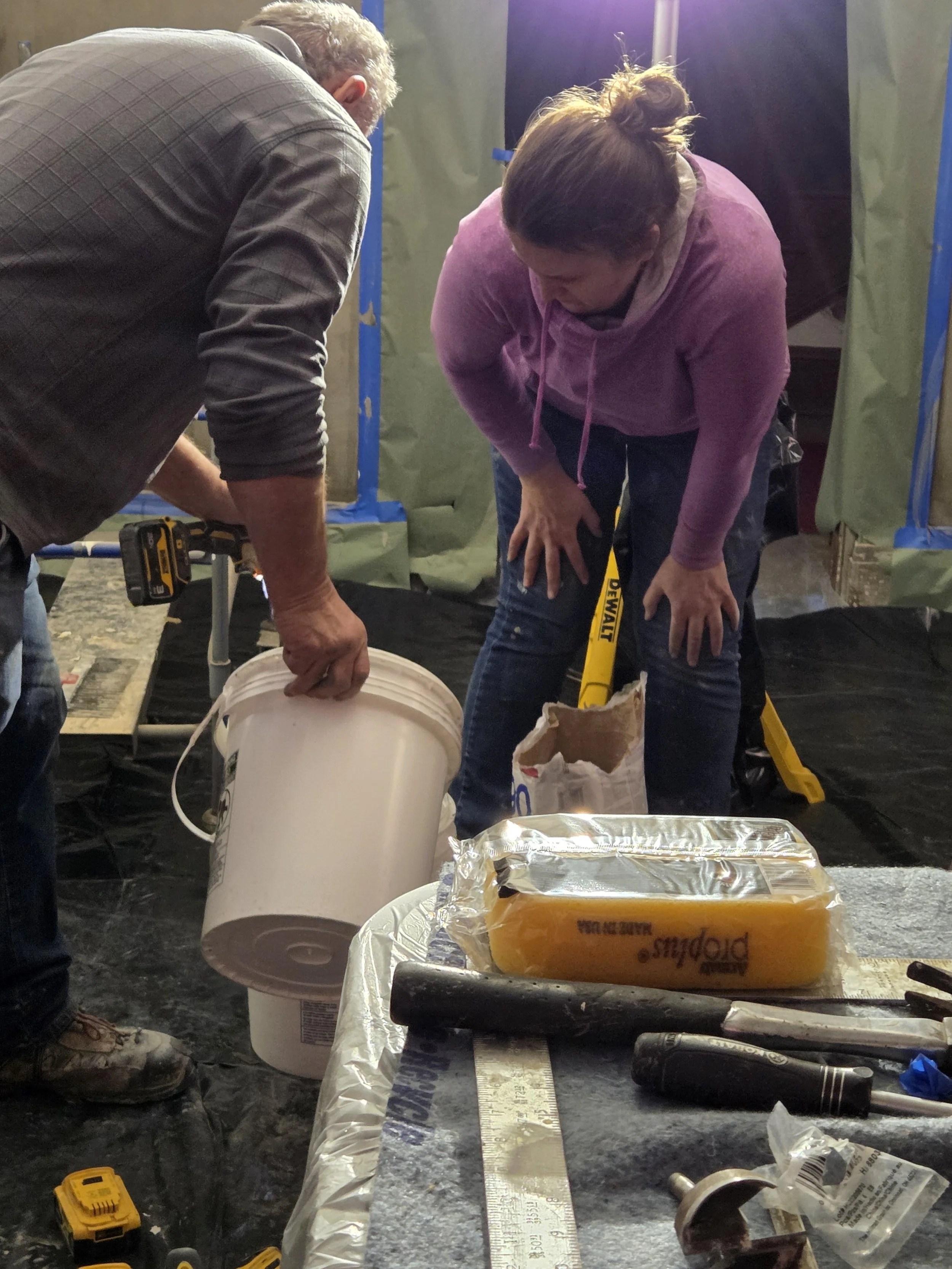 Two people working together on a construction or repair project, with one person pouring from a bucket and the other bent over, on a worksite with tools and materials.