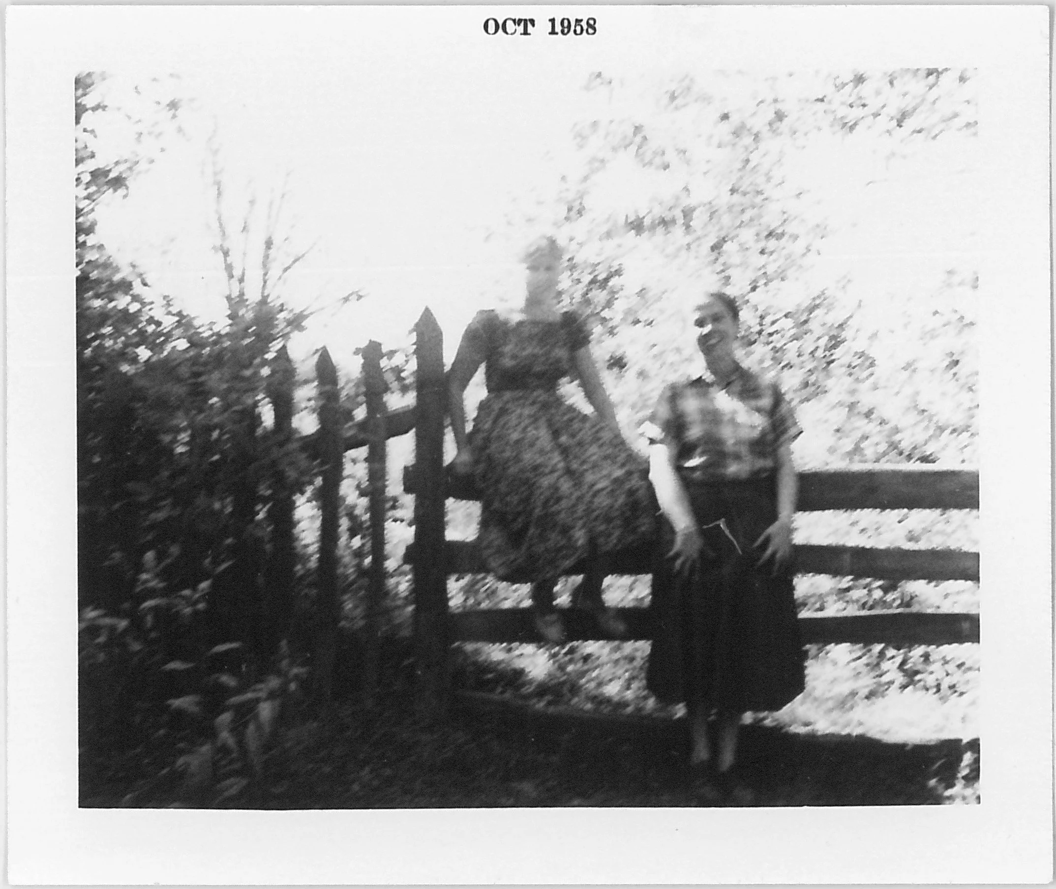 Black and white photo from October 1958 showing two women near a wooden fence, with one sitting on the fence and the other standing next to her and smiling.