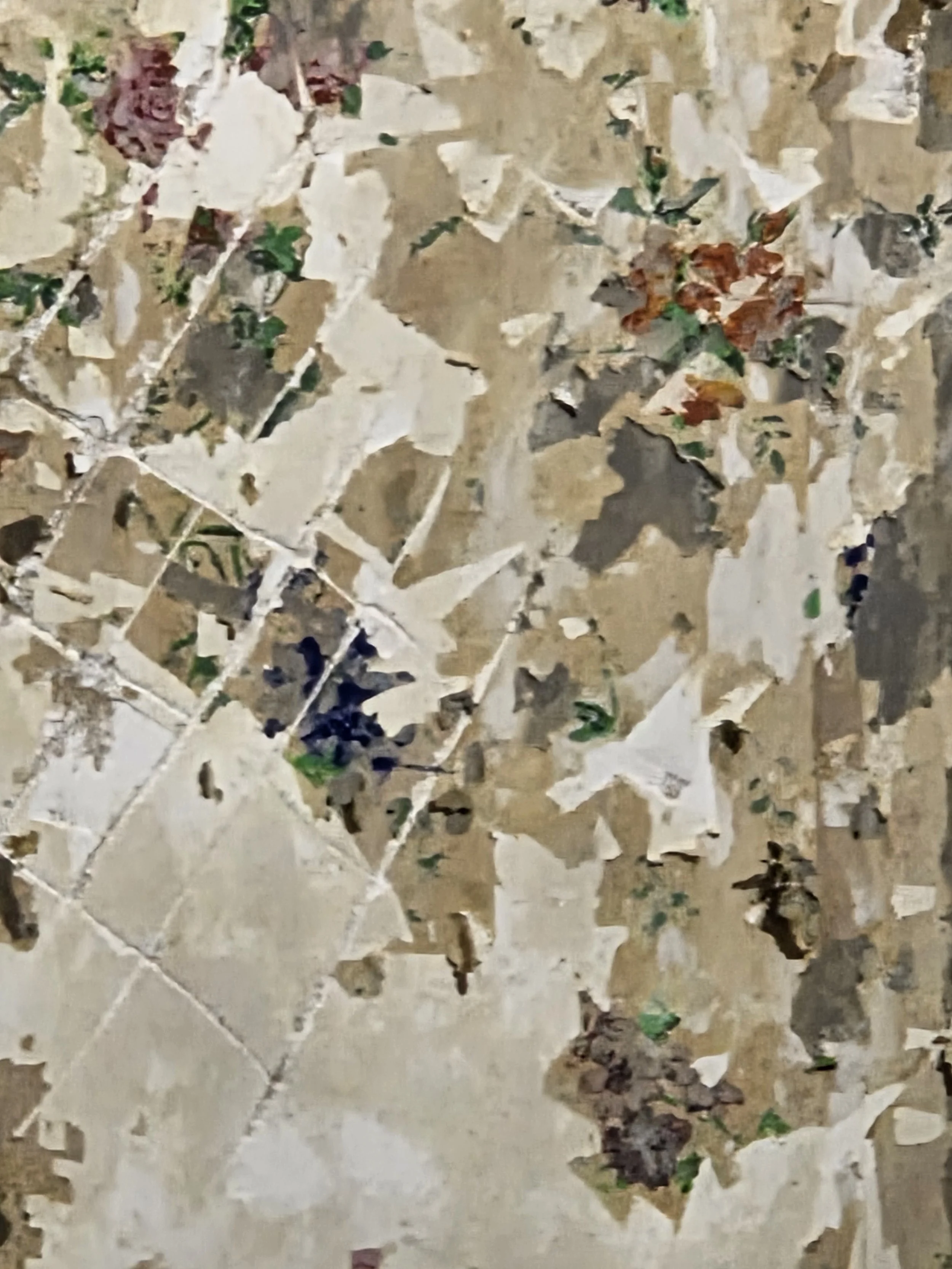 Close-up of a surface covered with torn and peeling pieces of wallpaper in various shades of beige, white, and brown, with small patches of green, purple, and reddish colors visible.