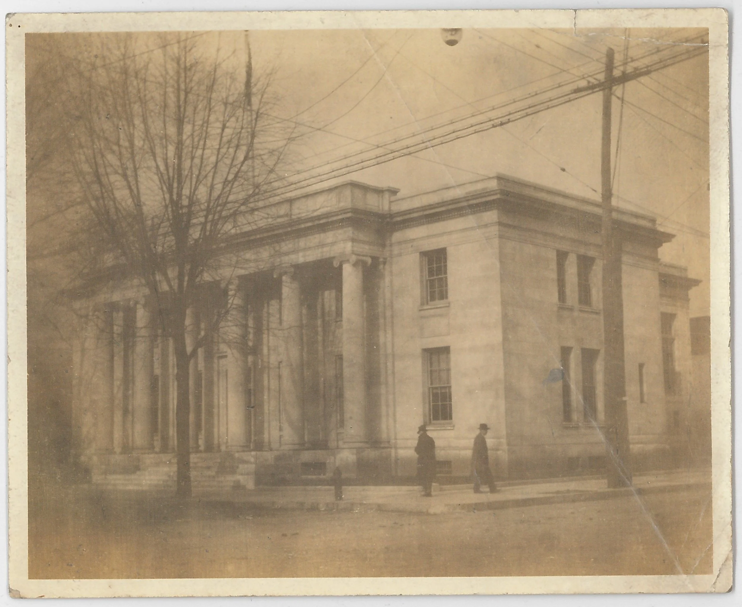 An old sepia-toned photograph of a stone building with classical architectural features such as columns and decorative moldings. Two men in hats are walking along the sidewalk in front of the building. There are power lines and a large leafless tree in the scene.