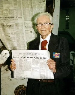 An elderly Sam Shaw with white hair and glasses, wearing a dark suit with a white shirt and red tie, holding the 100 year edition of the Moundsville Daily ECHO.