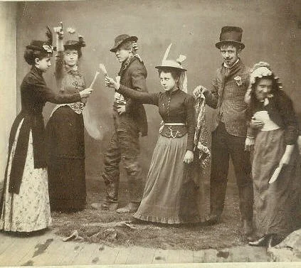 Group of six people dressed in 19th-century clothing, standing in a line, with some holding paper or objects, in an indoor setting.
