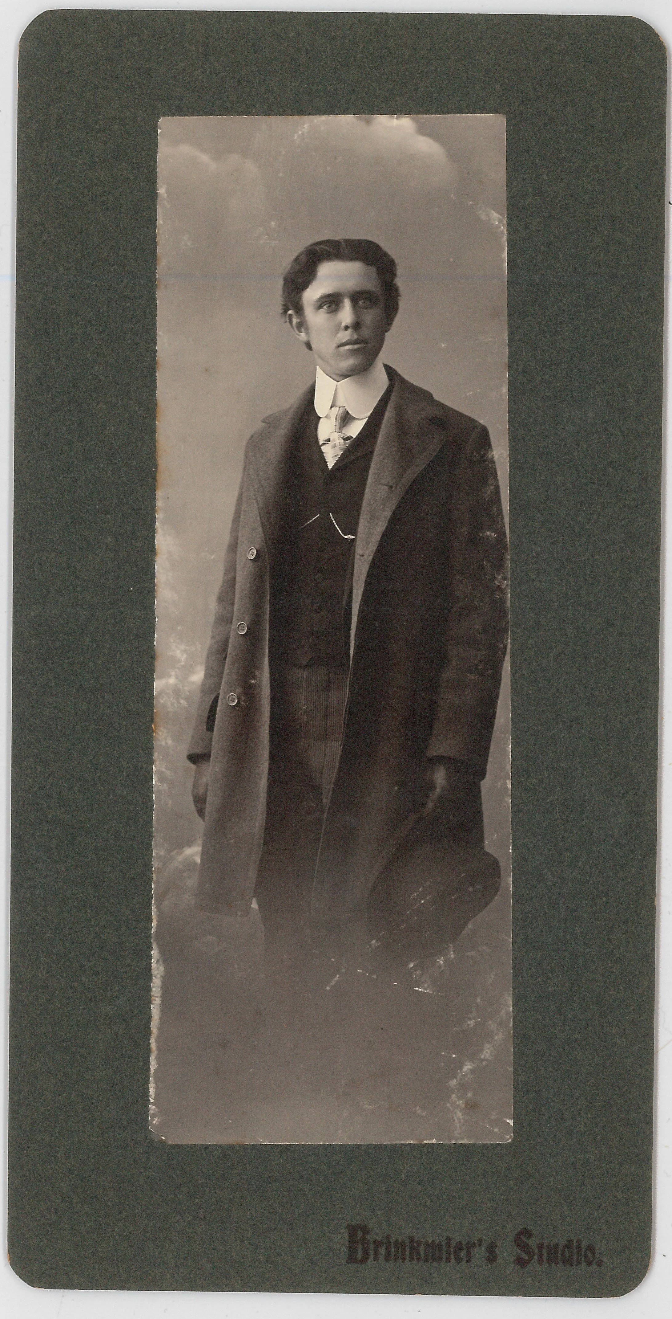 Black and white portrait of a young man in formal attire, standing outdoors with a cloudy sky background, encased in a cardboard frame that reads "Brooker's Studio."