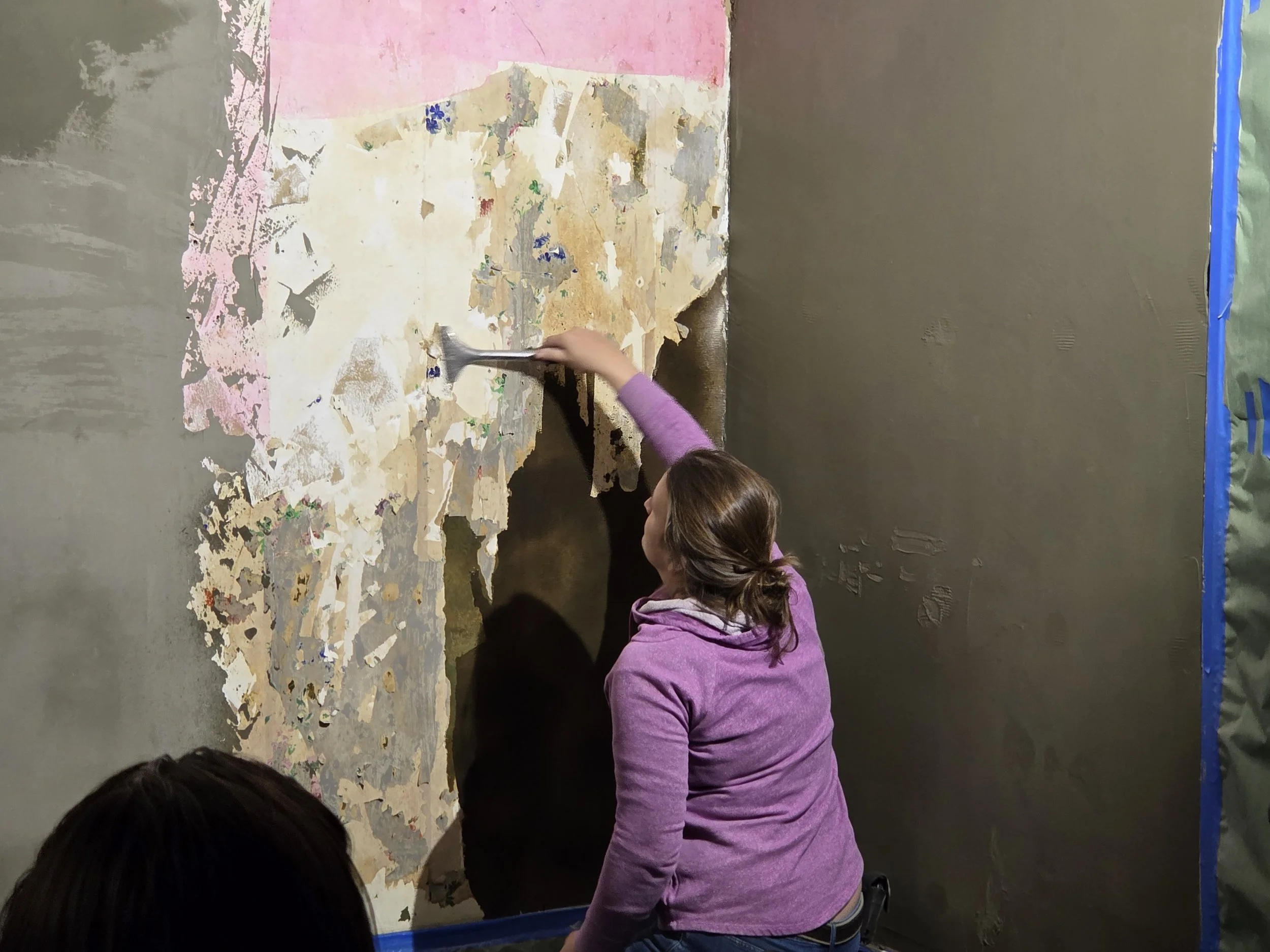 A woman in a purple hoodie removing old wallpaper from a wall with a scraper.