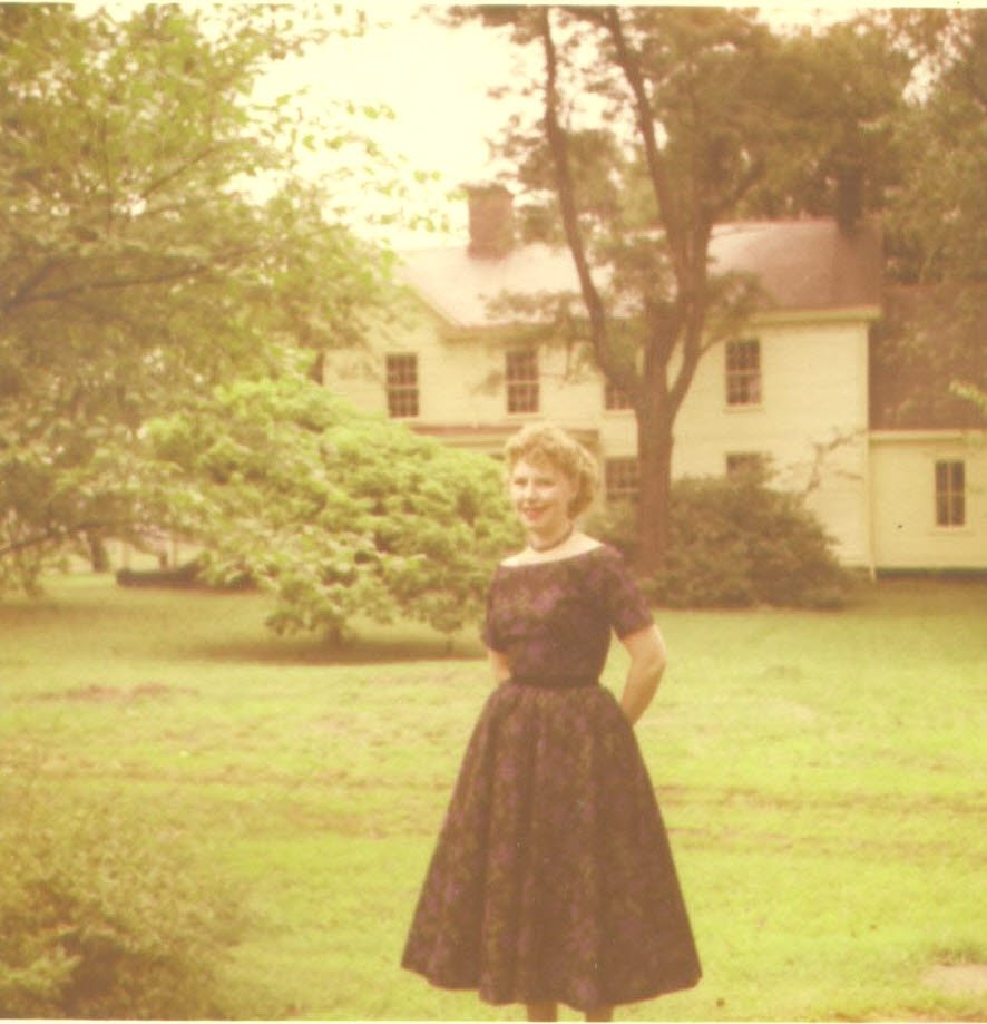 Lisa Cockayne with curly blonde hair standing in a grassy yard, wearing a dark floral dress with a white collar, smiling, with trees and a large white house in the background.