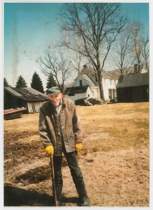An elderly man standing outdoors on a patchy grassy area, holding a shovel, wearing a winter coat, gloves, and a hat, with leafless trees and a house in the background.
