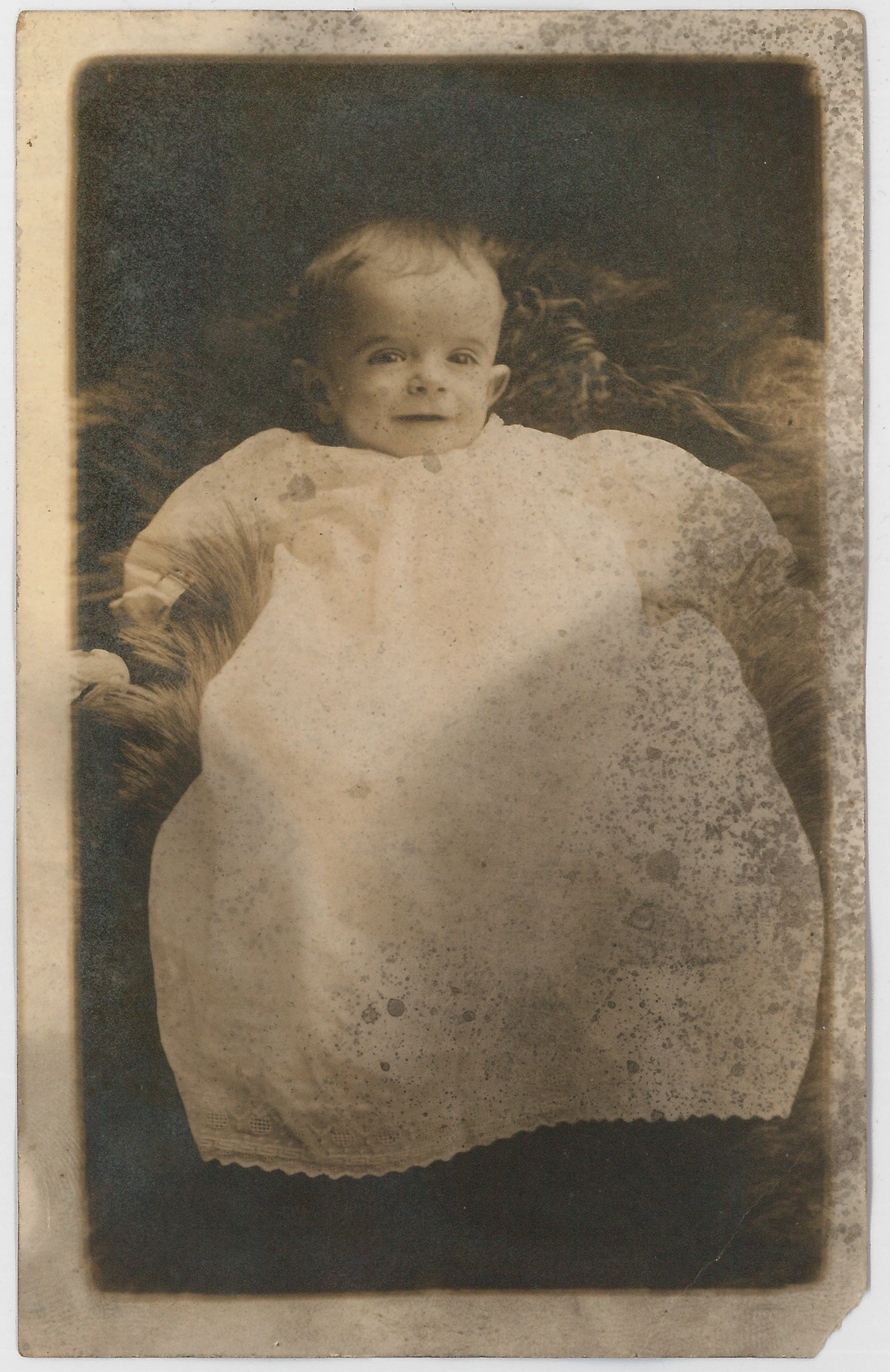 Black and white photo of a young child sitting in a wicker chair, smiling, with a dark background.