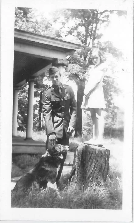 A black and white photo of a man in a military uniform standing outdoors next to a dog, a girl standing on a tree stump, and a house with a porch in the background, surrounded by trees.