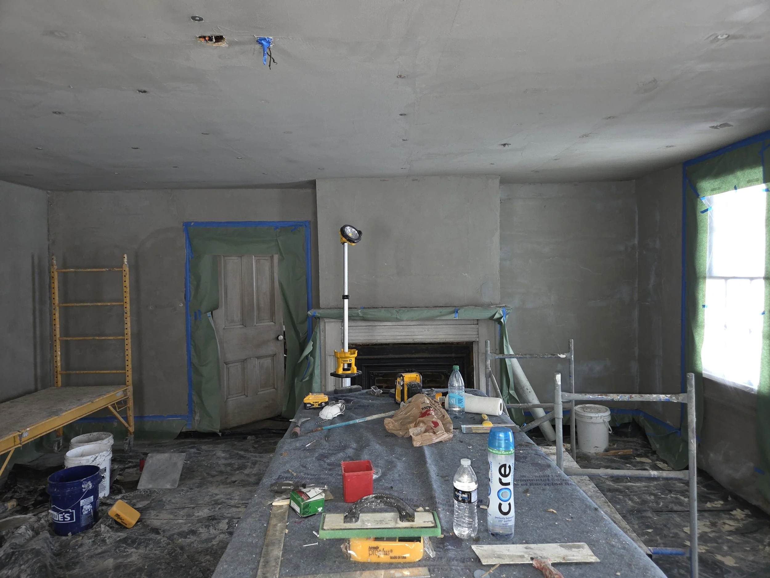 Room under construction with scaffolding, construction tools, supplies, and taped window and door coverings.