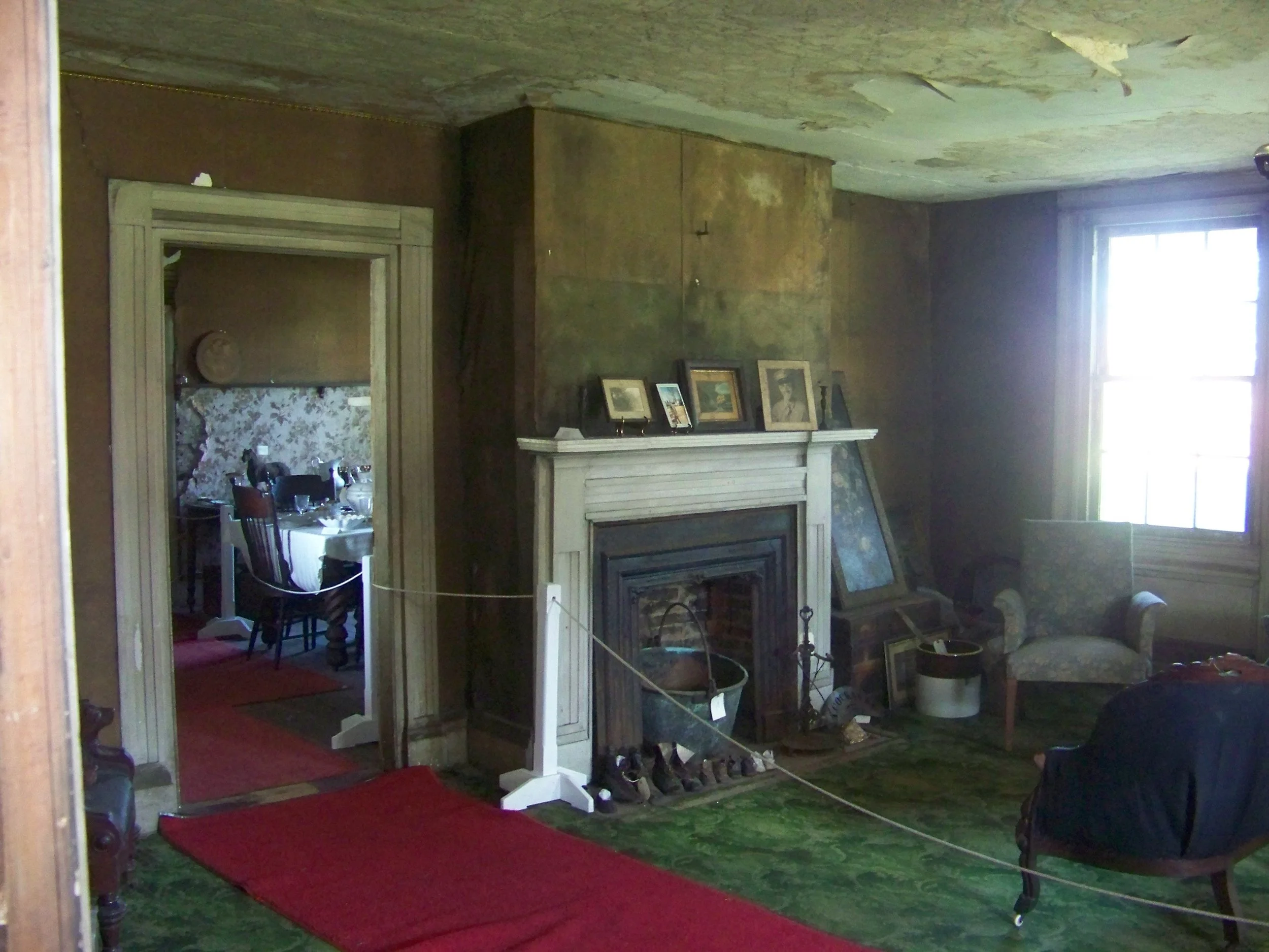 An old living room with a green carpet, red runner, fireplace with framed pictures on the mantel, and a single armchair near a window.