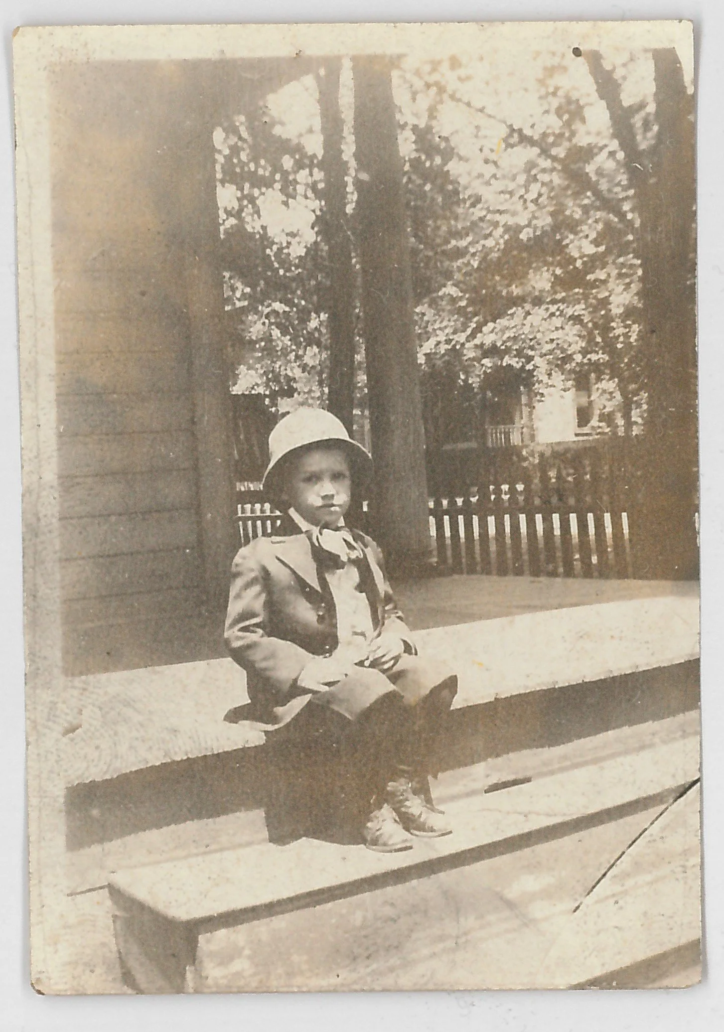 A young boy dressed in vintage clothing, including a suit and military-style hat, sitting on a park bench with trees and houses in the background.